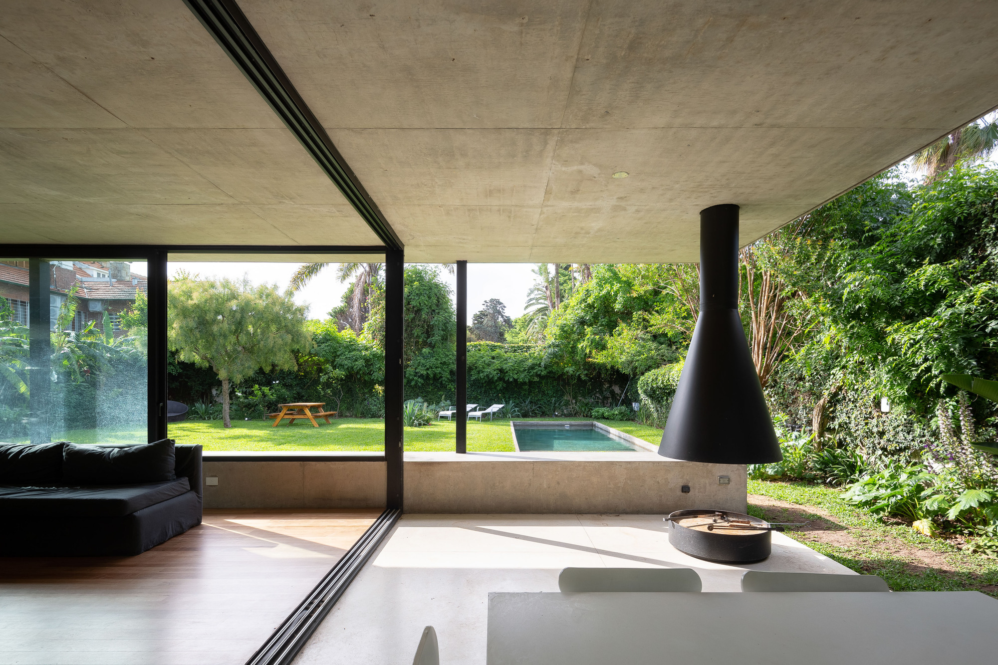 Living space opening through sliding glass to a lawn and pool, with a black hanging fireplace under a concrete ceiling