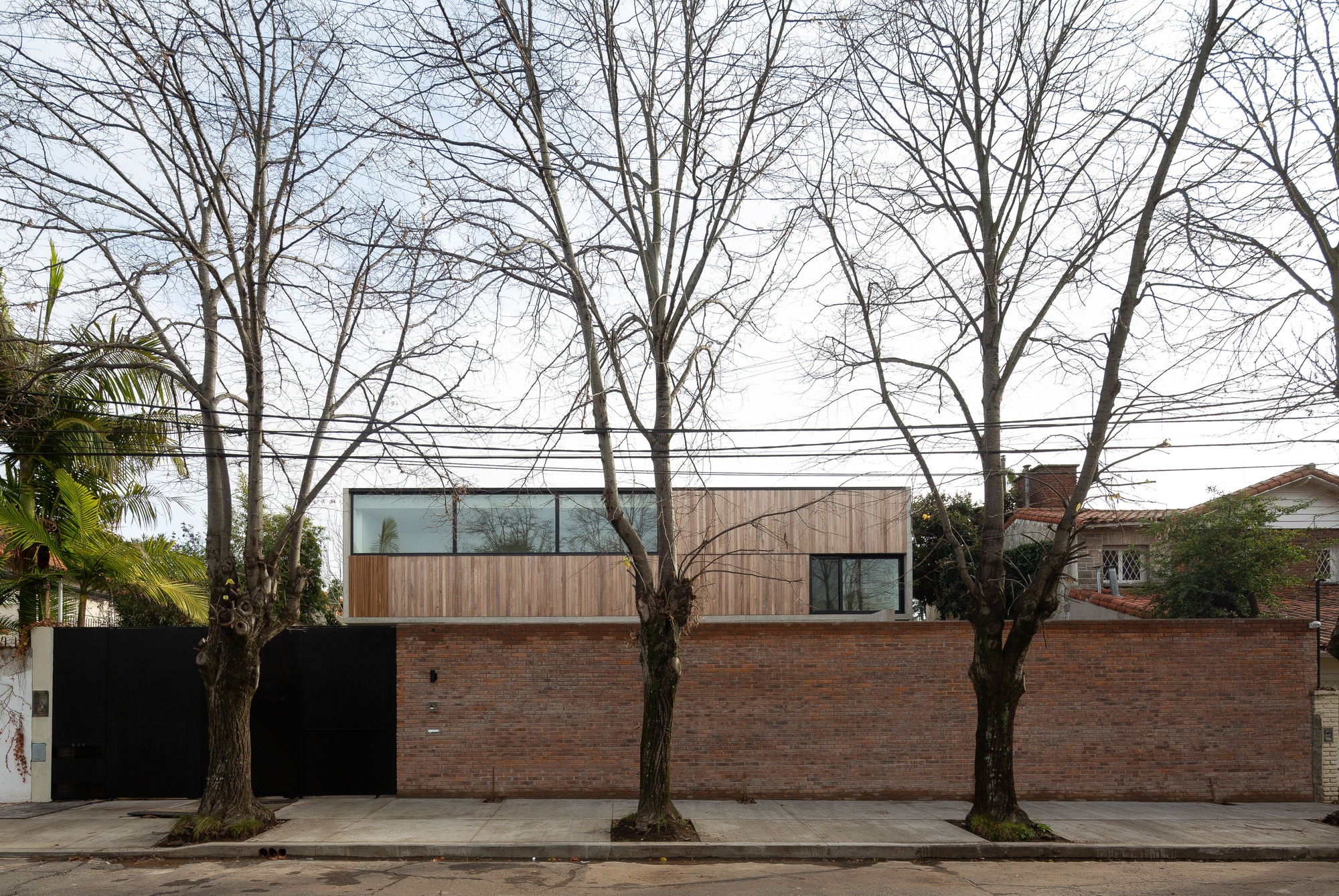 Street elevation of a brick wall and black gate with a timber-clad upper floor, seen through bare winter trees