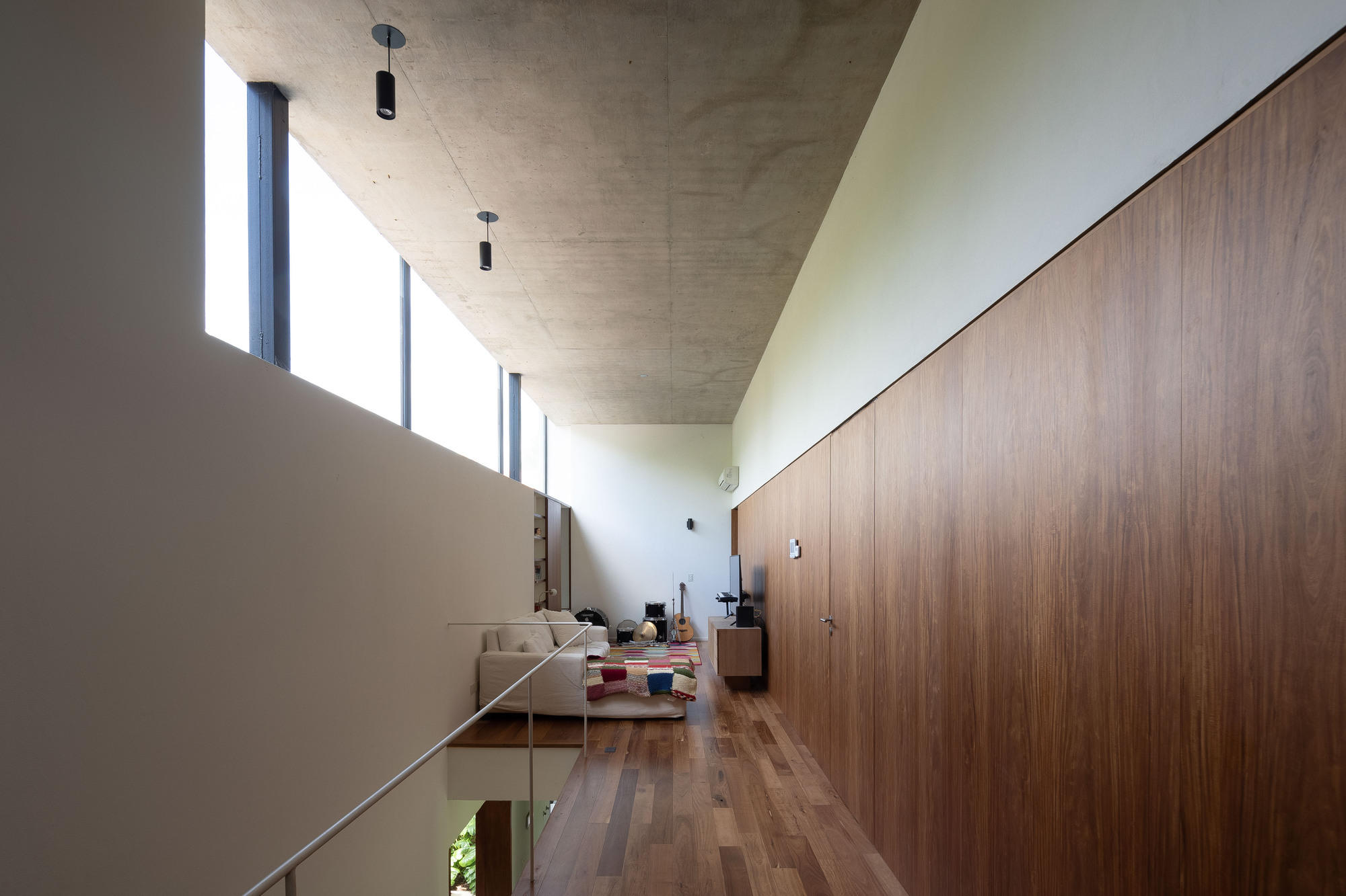 Upper hallway with a long walnut panel wall, board-formed concrete ceiling and a clerestory window washing light over a lounge at the far end