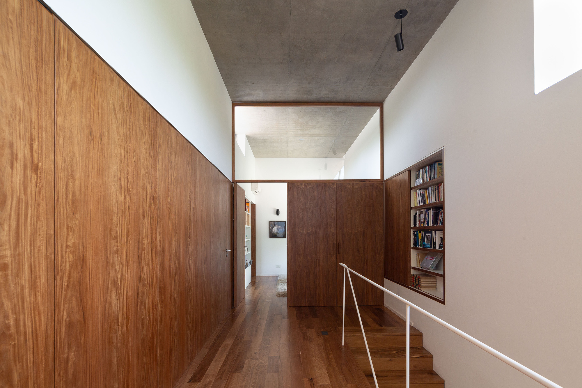 Corridor beside a timber-clad wall with a recessed bookshelf, white handrail and stair descending beneath a concrete ceiling and interior glazed opening