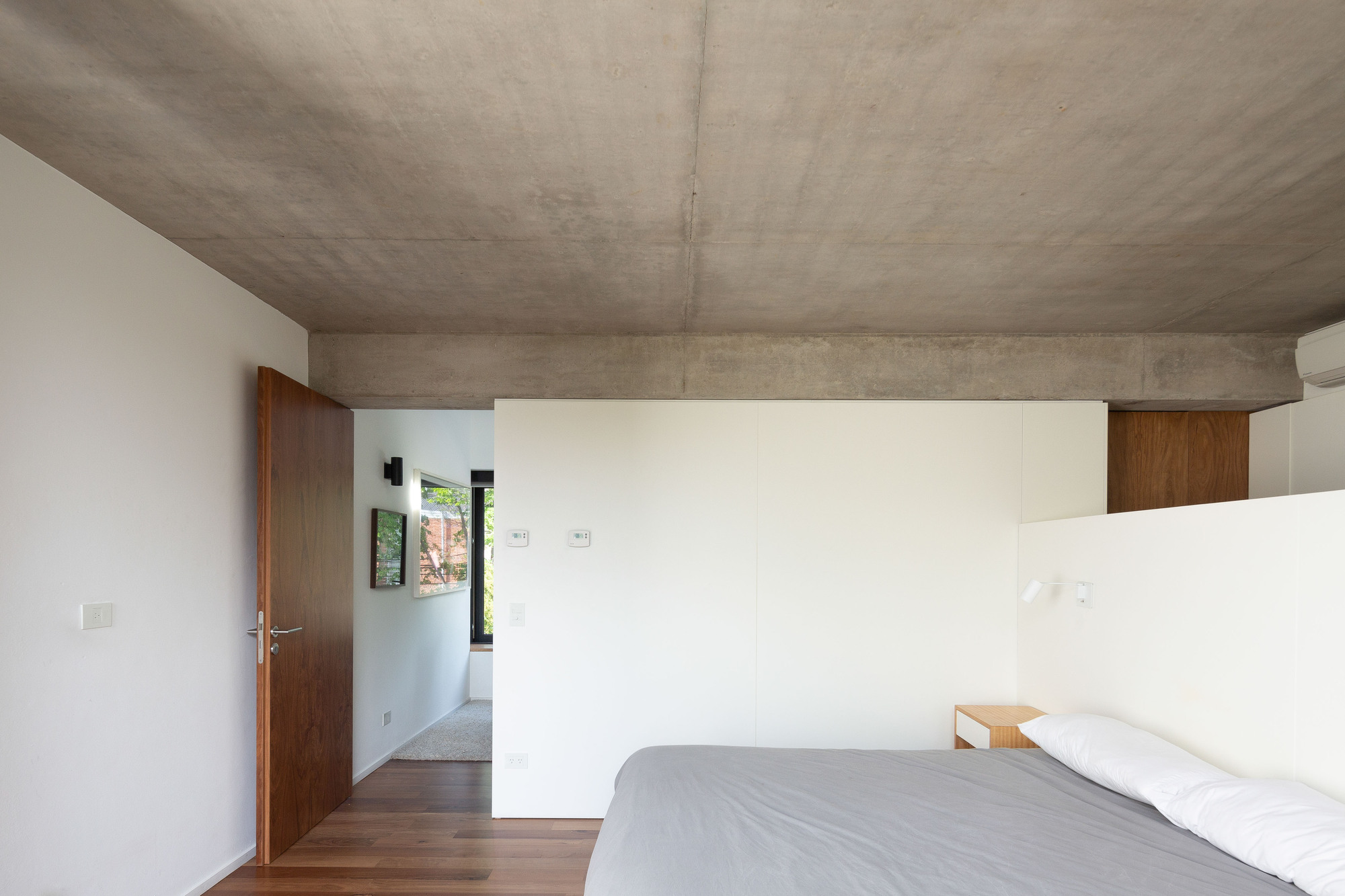 Bedroom with exposed concrete ceiling and beam, a walnut door standing open onto a bright passage with a framed window