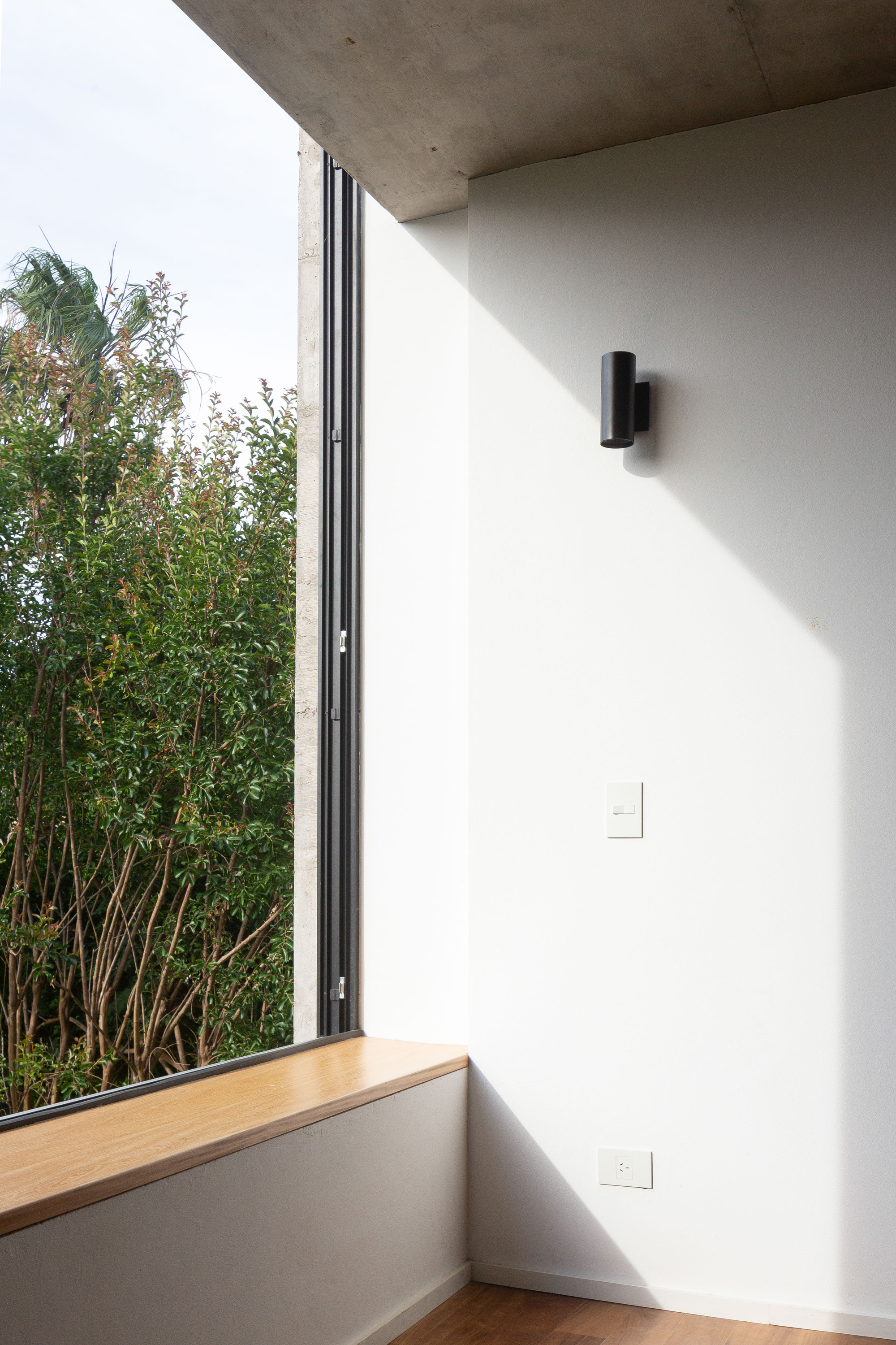 Corner window with a timber sill framing a leafy hedge, a black cylinder wall light catching a diagonal band of sunlight