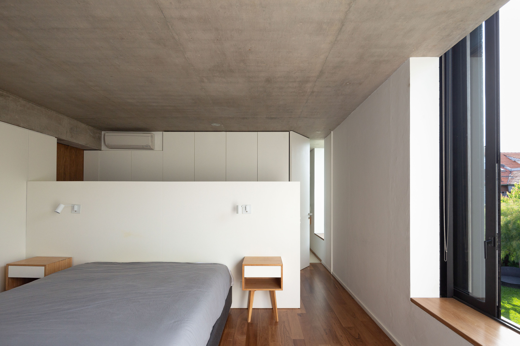 Bedroom with a low white headboard partition, wooden bedside tables and a tall black-framed window opening toward a green lawn and tiled roof