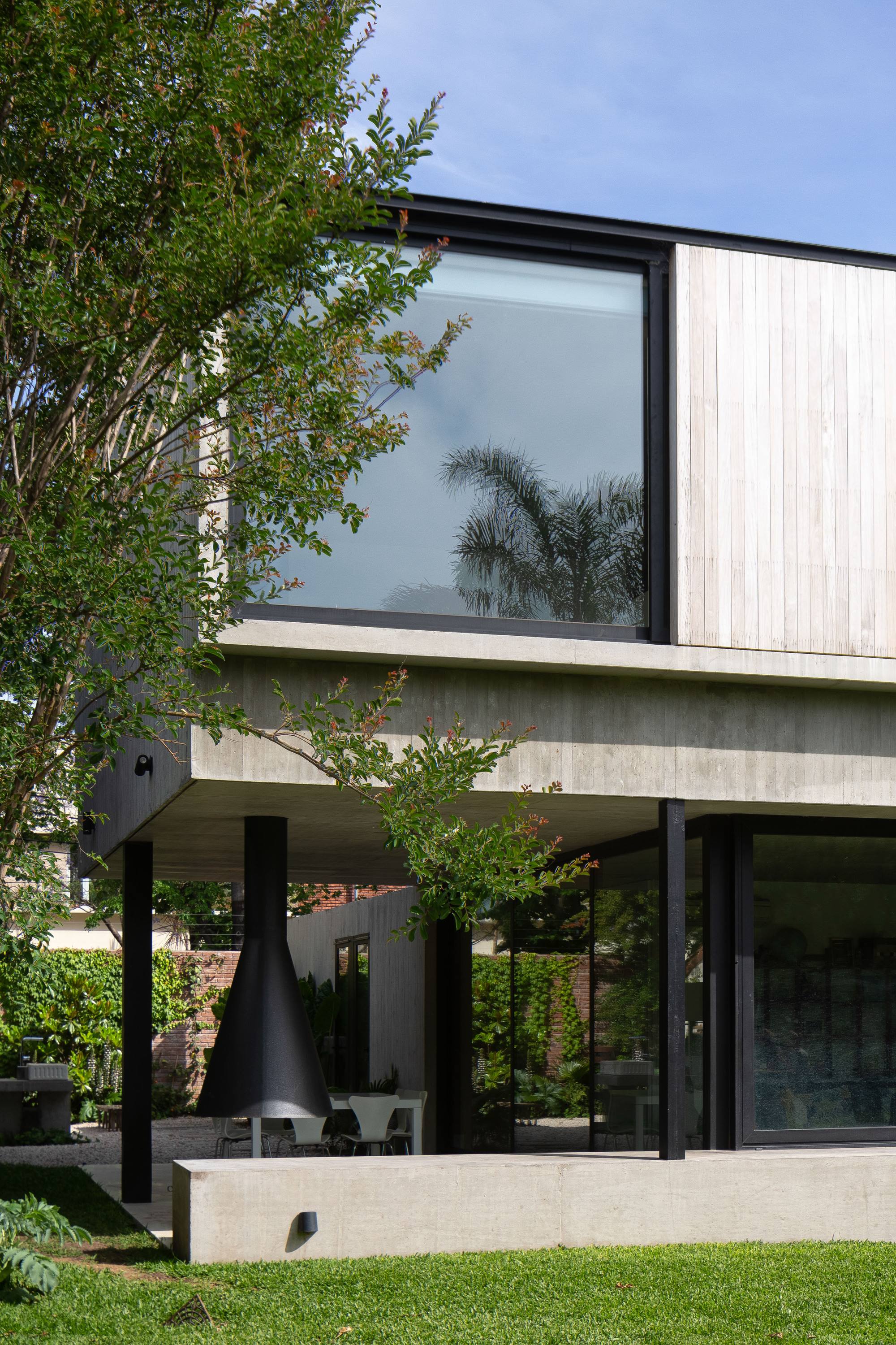 Cantilevered concrete upper volume with timber cladding and a black conical fireplace on the terrace below, under a blue sky