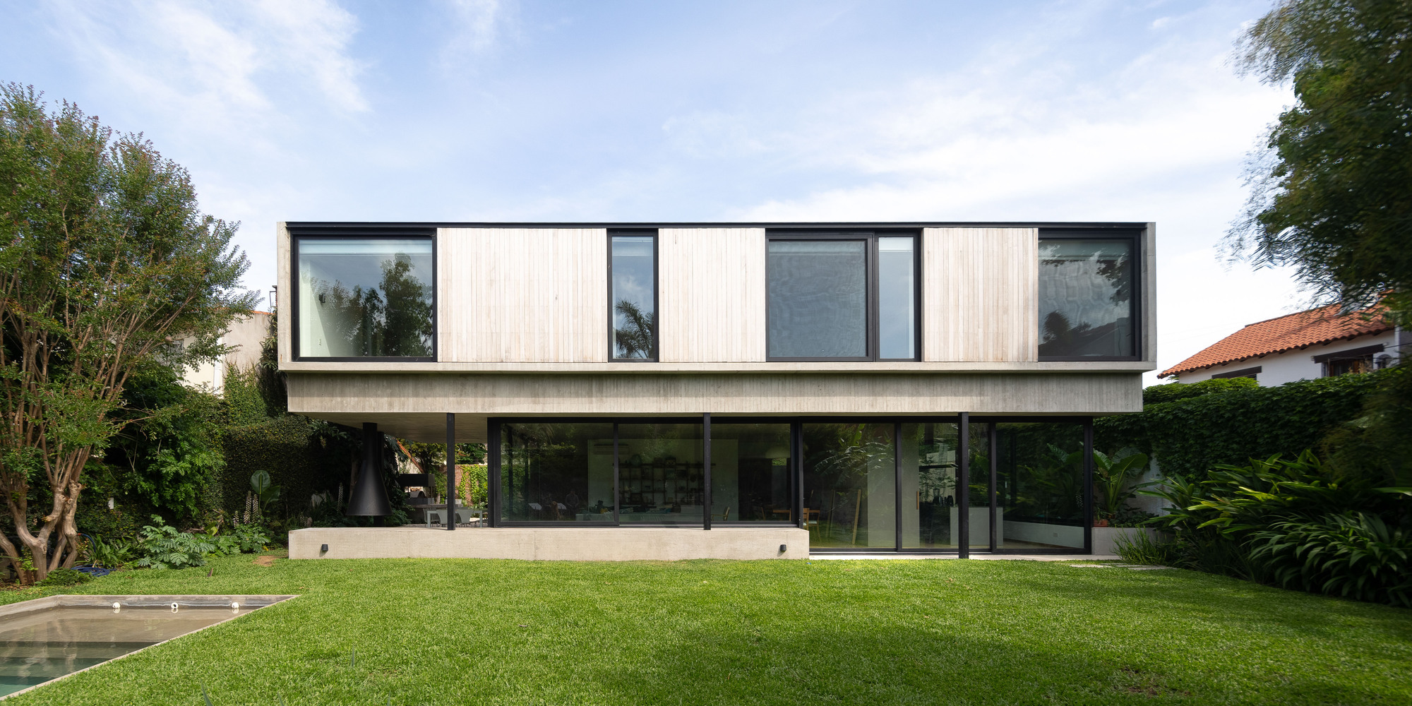 Rear elevation of the two-storey house with a timber-clad box over a glazed ground floor, facing a lawn and pool edge