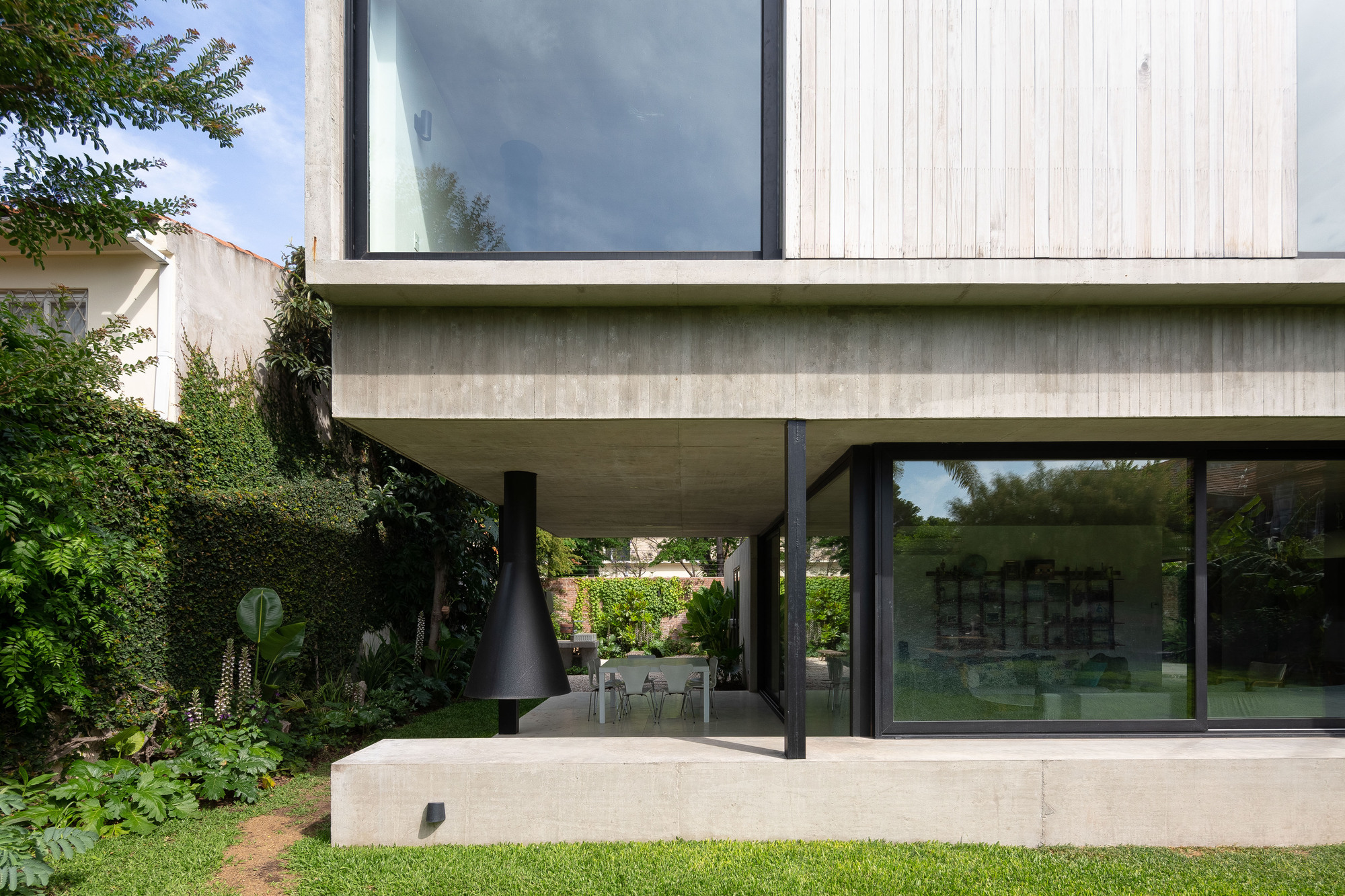 Covered concrete terrace with a hanging black fireplace and dining table beside tall glass sliding doors, hedges and garden plants at left