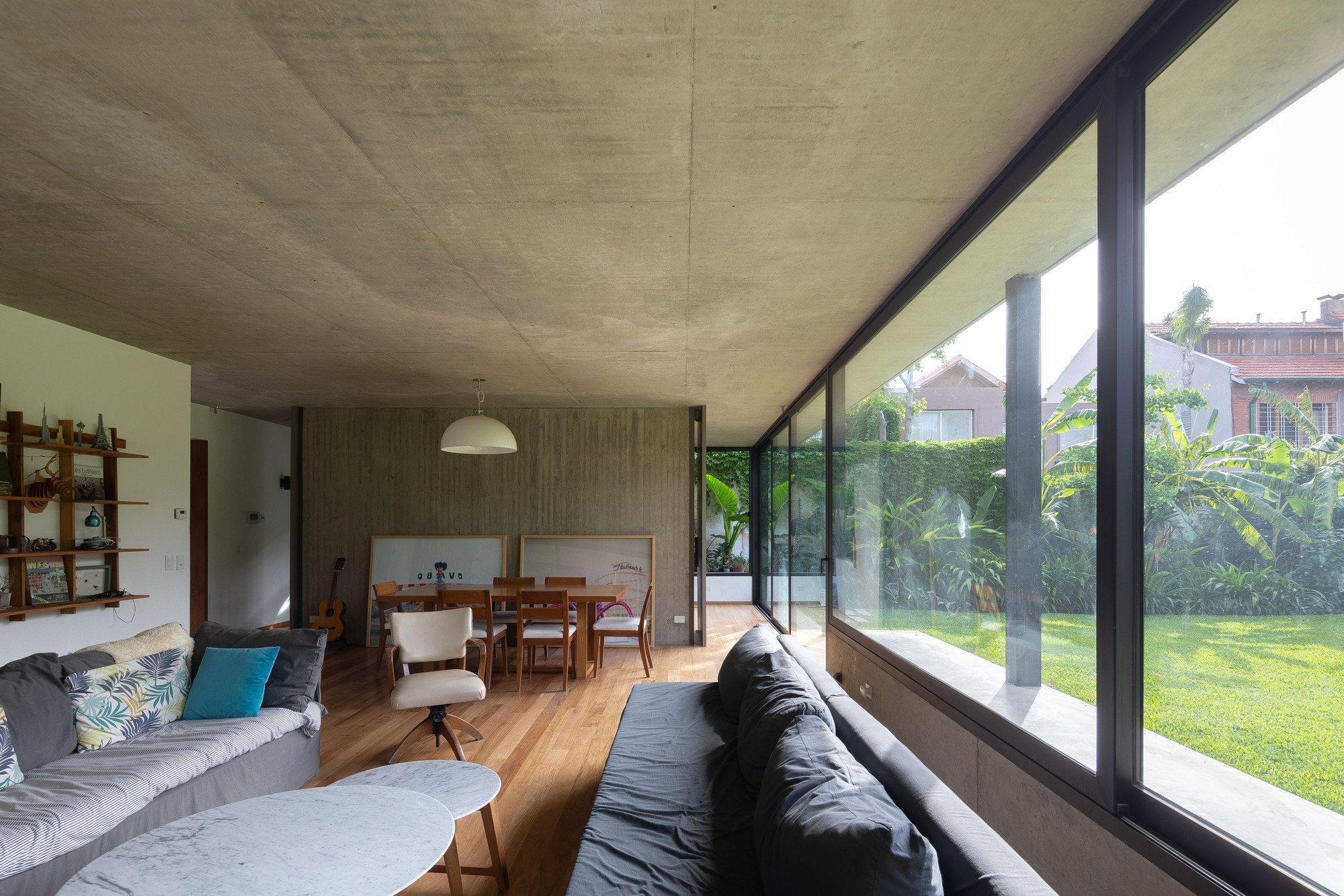 Living room with a concrete ceiling, timber floor and long window wall opening onto a sunlit lawn and tropical plants