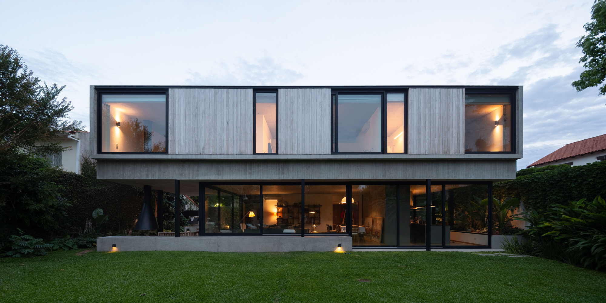 Rear elevation of the two-storey house with a board-formed concrete upper volume over a glazed ground floor, lit at dusk across the lawn