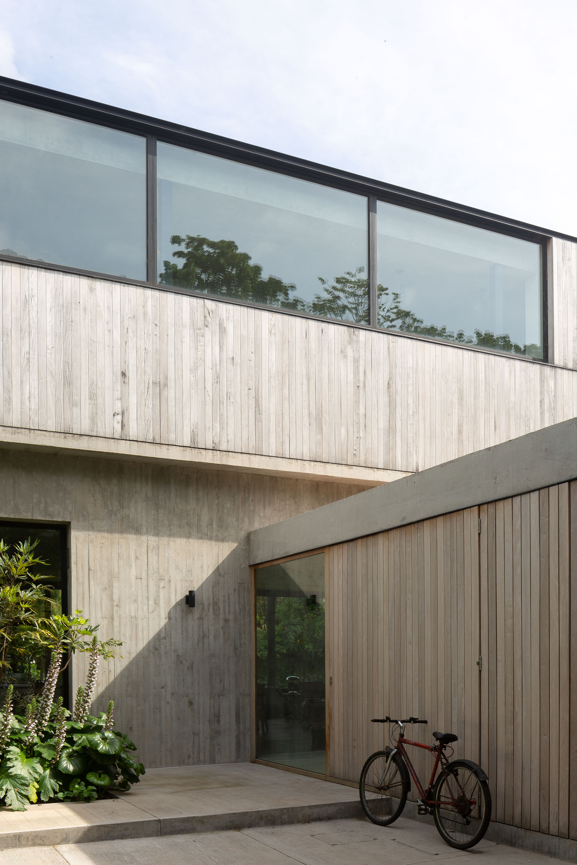 Entry courtyard corner with vertical timber cladding, board-marked concrete walls and a red bicycle parked beside a planted bed in daylight