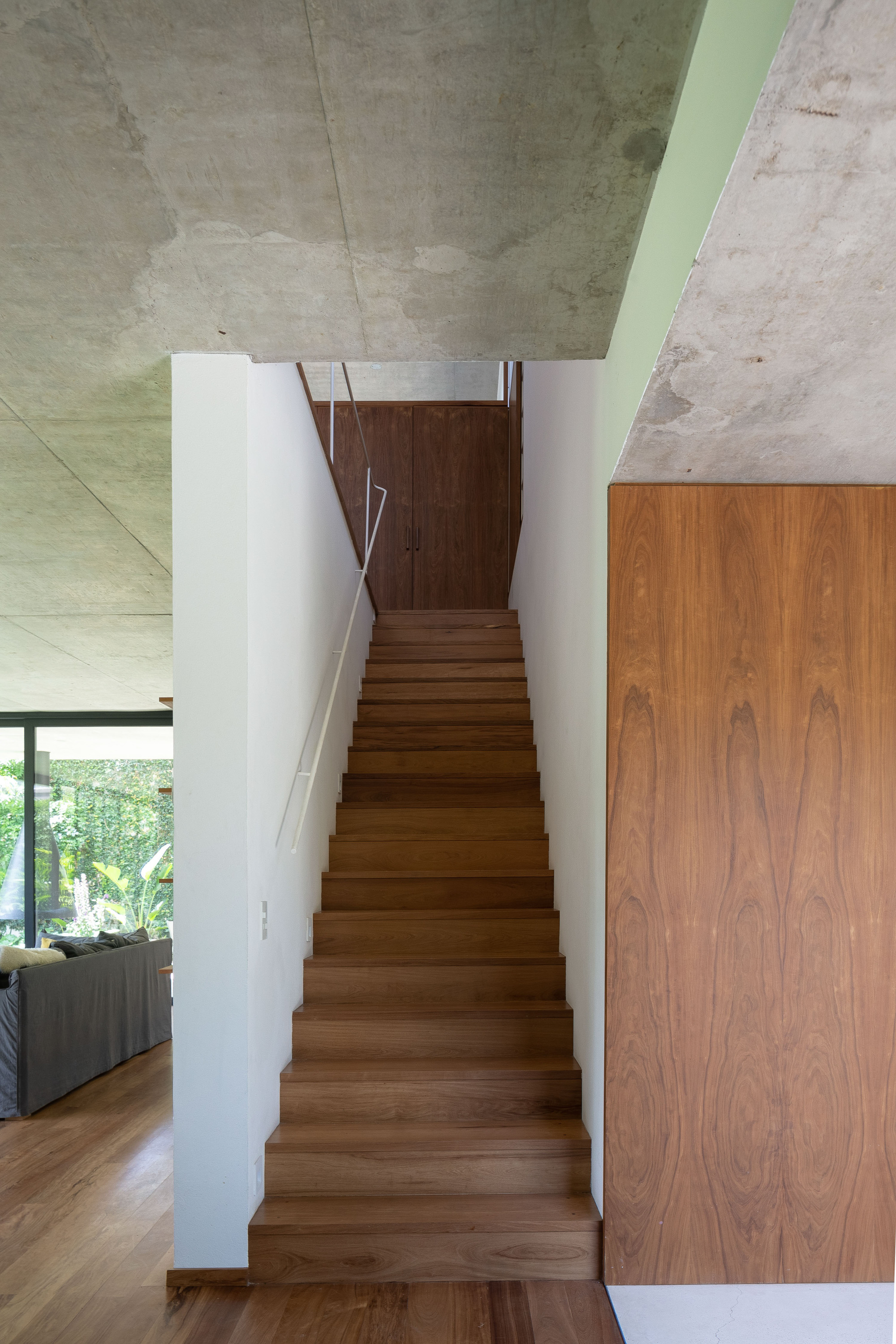Straight timber staircase between a white wall and a walnut-veneer panel, under a raw concrete ceiling with a living room glimpsed at left