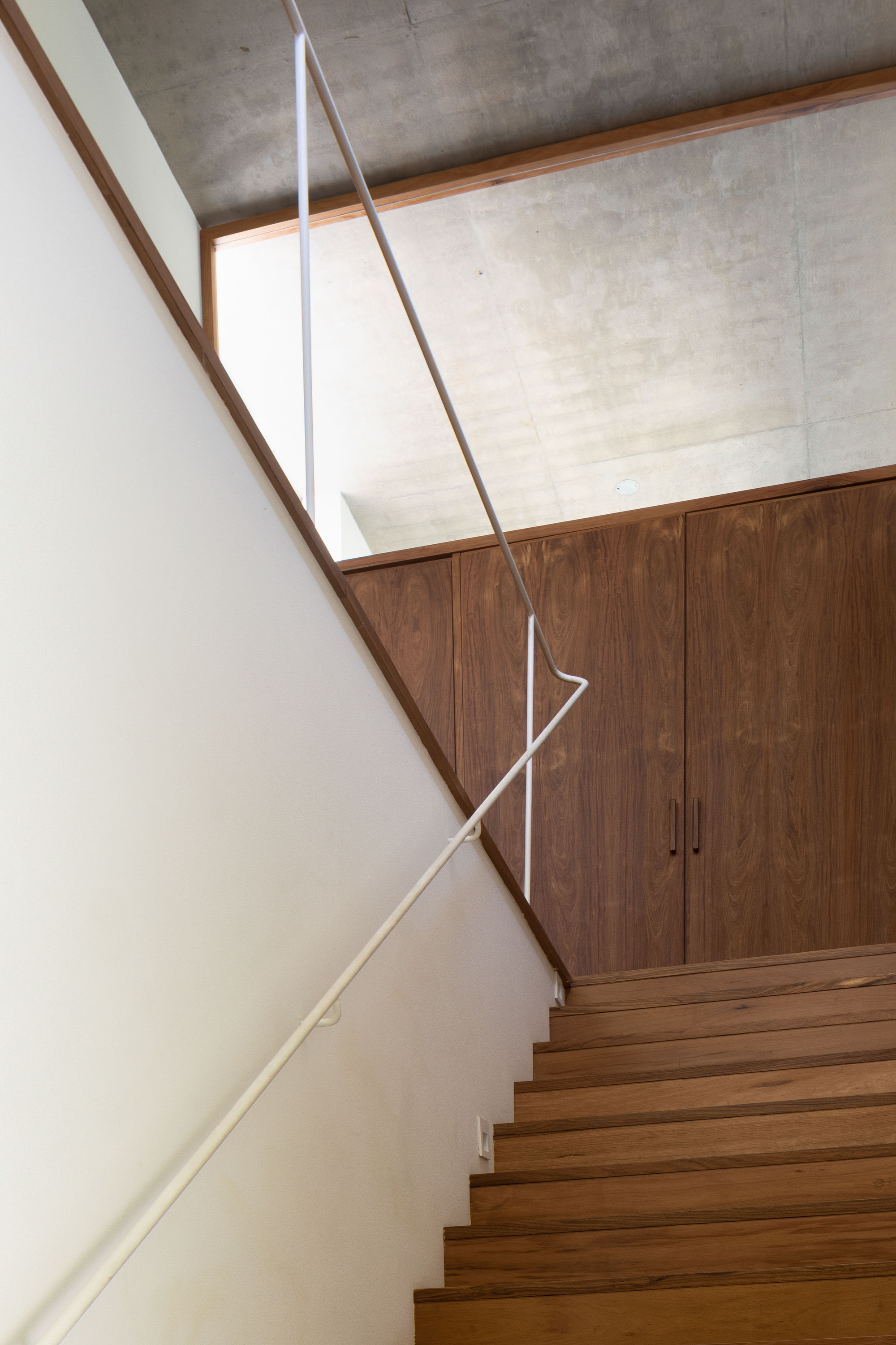 Looking up the timber stair past a white handrail to walnut cabinet doors and a concrete ceiling lit by a high window