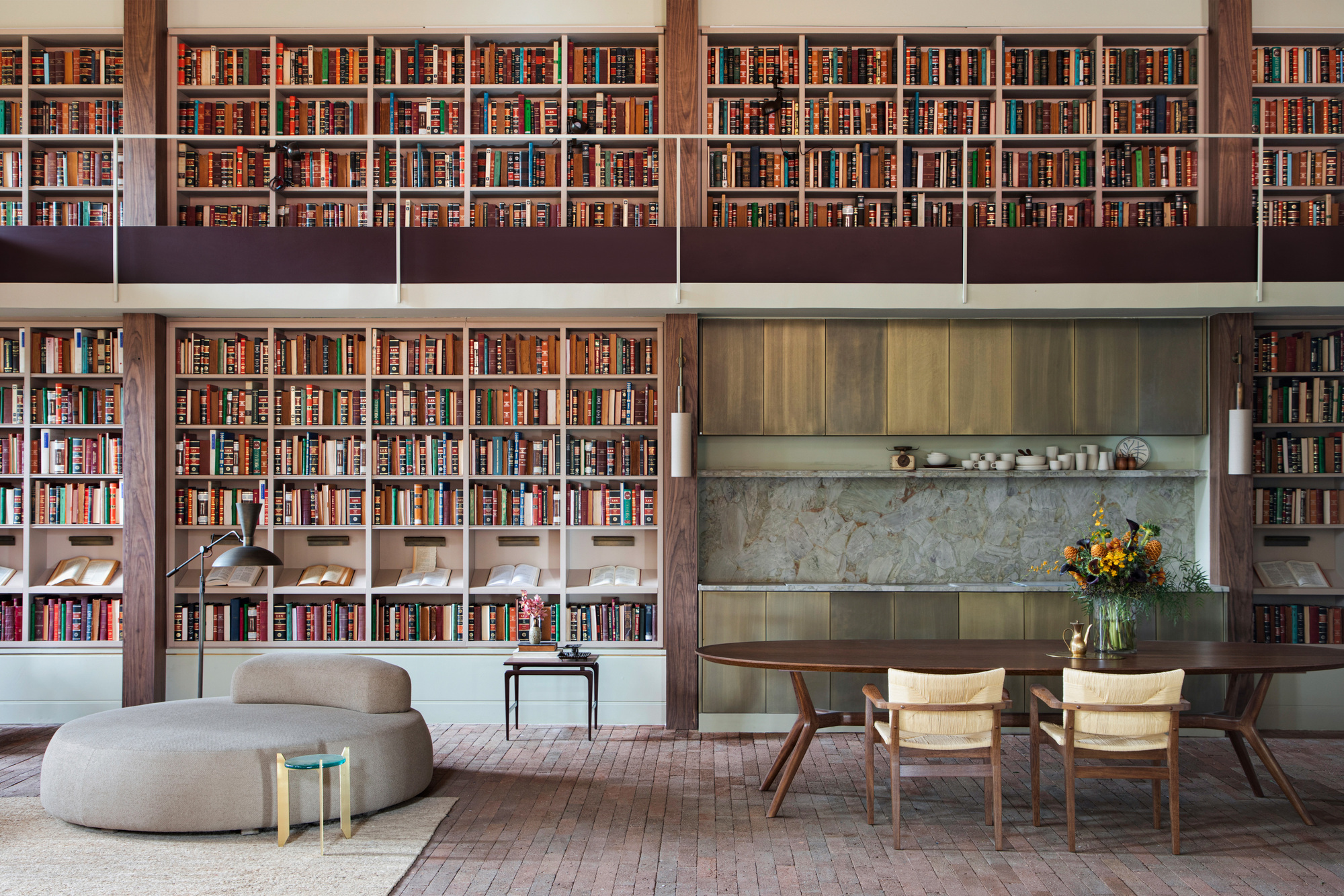 Double-height library wall of books with a mezzanine rail above a dining table, stone backsplash and a vase of orange flowers