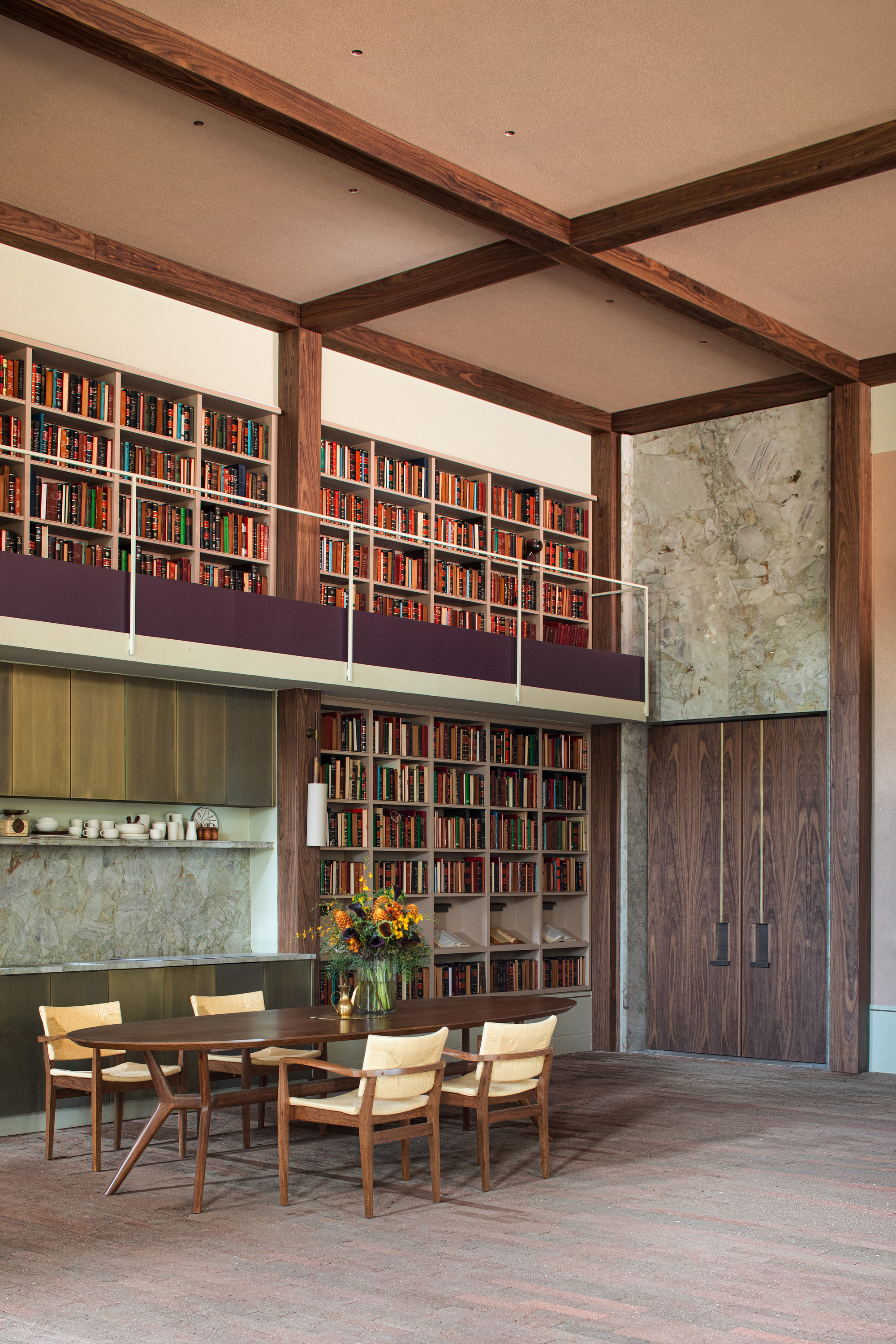 Oval dining table with cream chairs below a bookshelf mezzanine, beside tall walnut doors and a veined stone wall