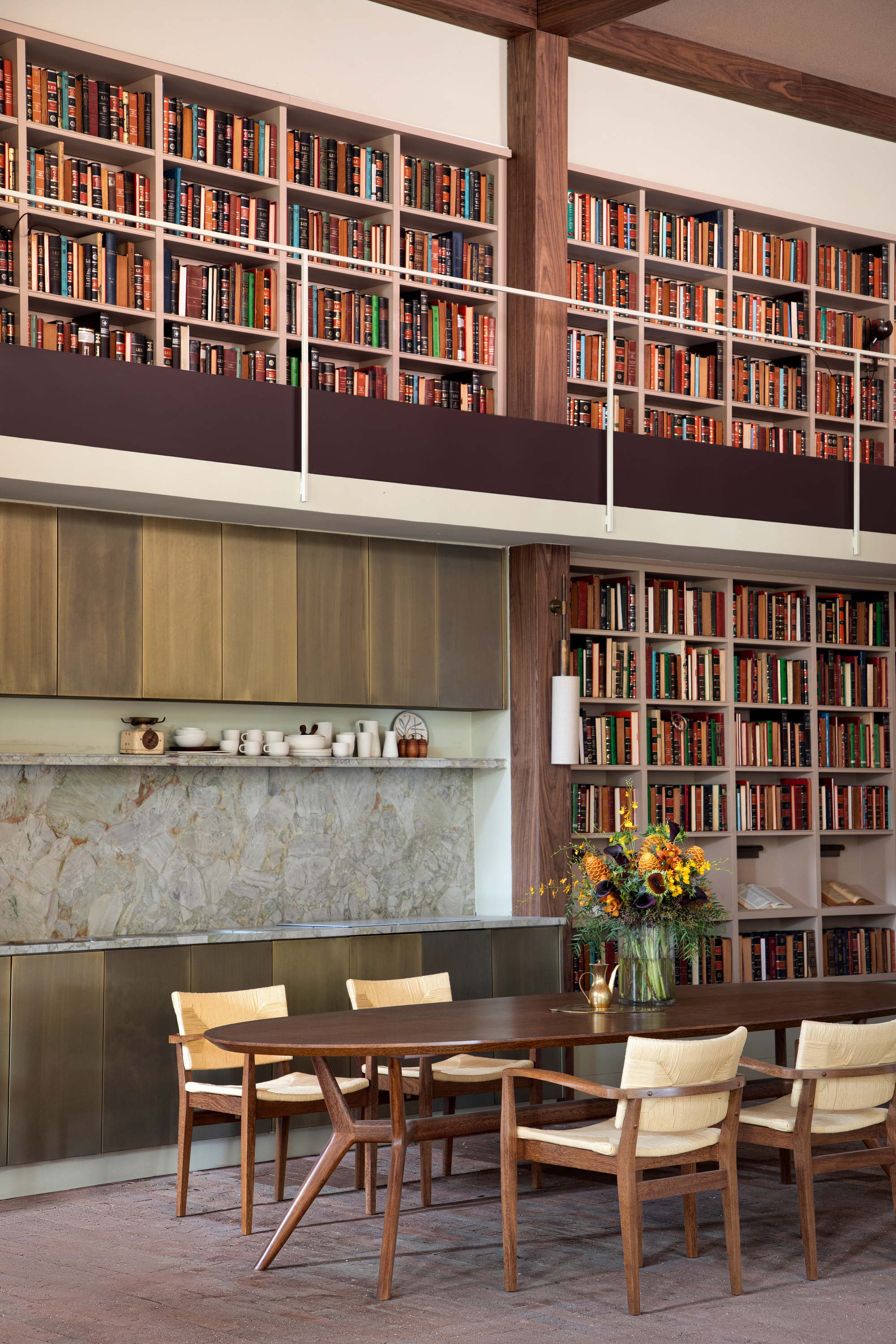 Oval walnut dining table and cane chairs below a double-height library gallery, with a vase of sunflowers and a stone backsplash