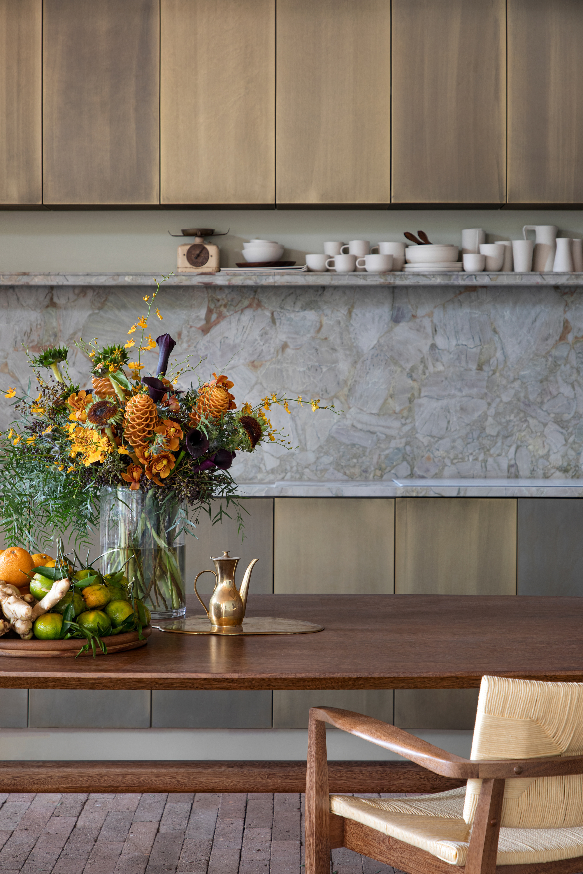 Kitchen wall of veined stone backsplash and open shelf with white ceramics above a timber table, flower arrangement and citrus bowl
