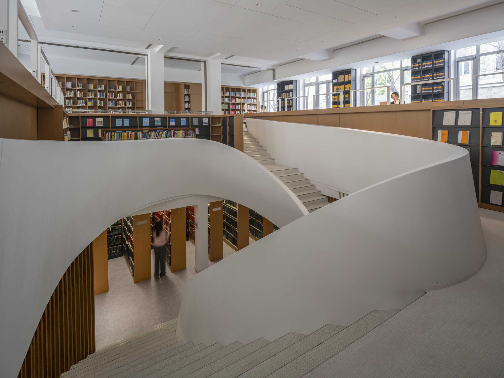 Curving white stair balustrades sweeping over library stacks, with a reader standing between timber bookshelves in daylight from tall windows