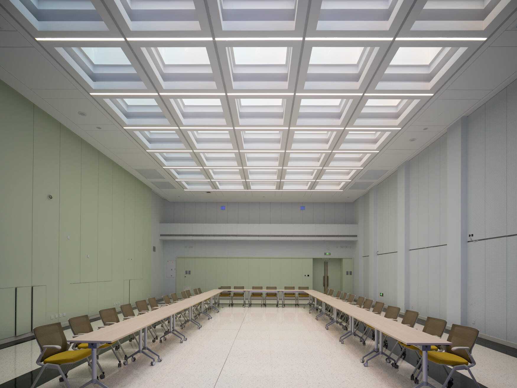 Meeting room with a coffered skylight ceiling above U-shaped tables and yellow-cushioned chairs on a terrazzo floor