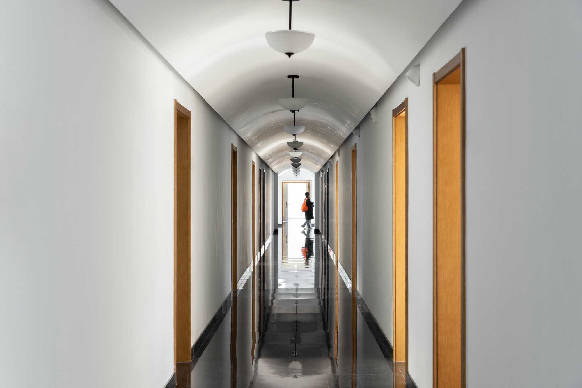 Barrel-vaulted corridor with pendant lamps and timber door frames, a person in an orange backpack reflected in the glossy dark floor