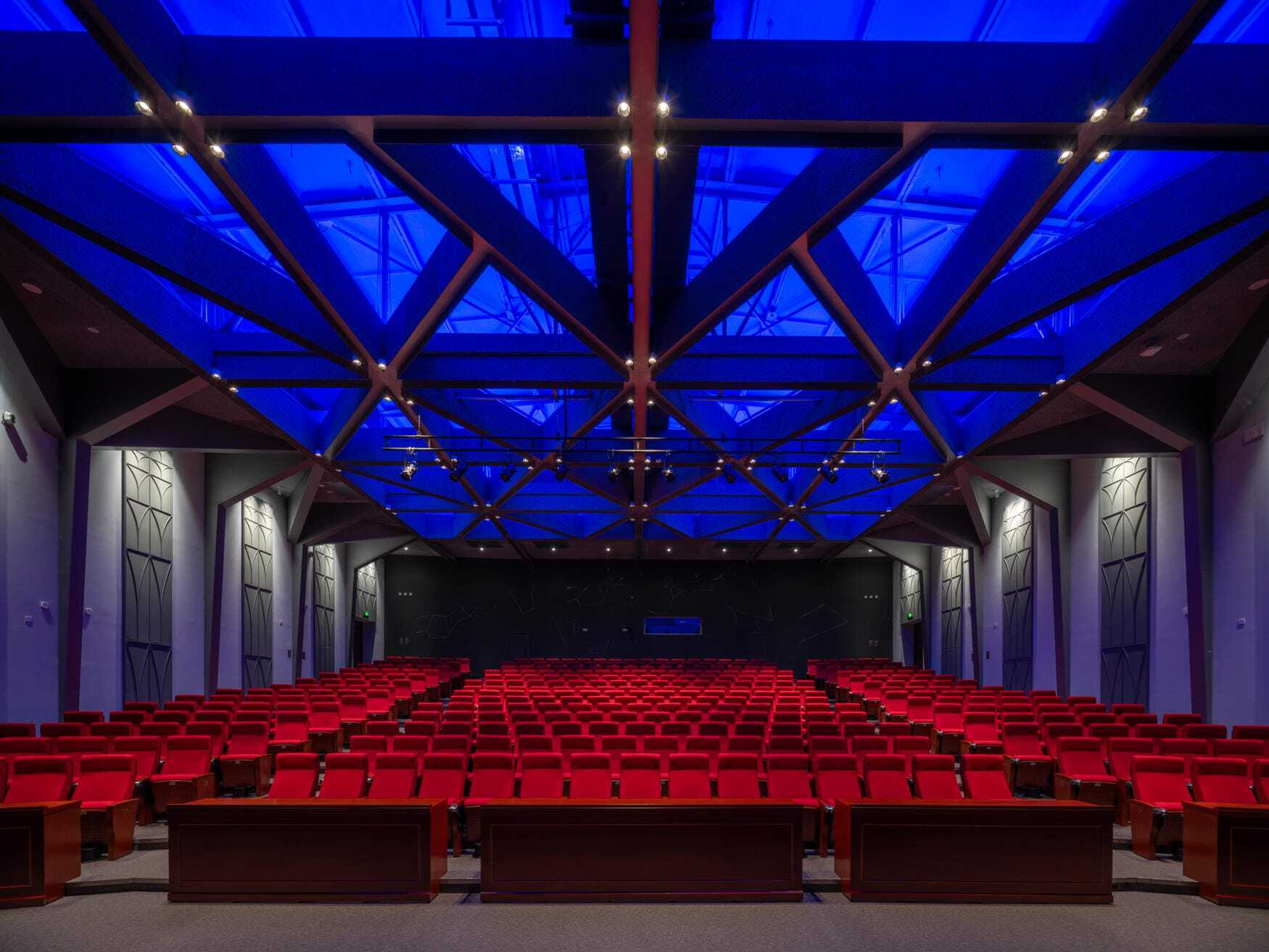 Auditorium with red seats facing a stage, lit by blue light through a steel truss roof and skylights