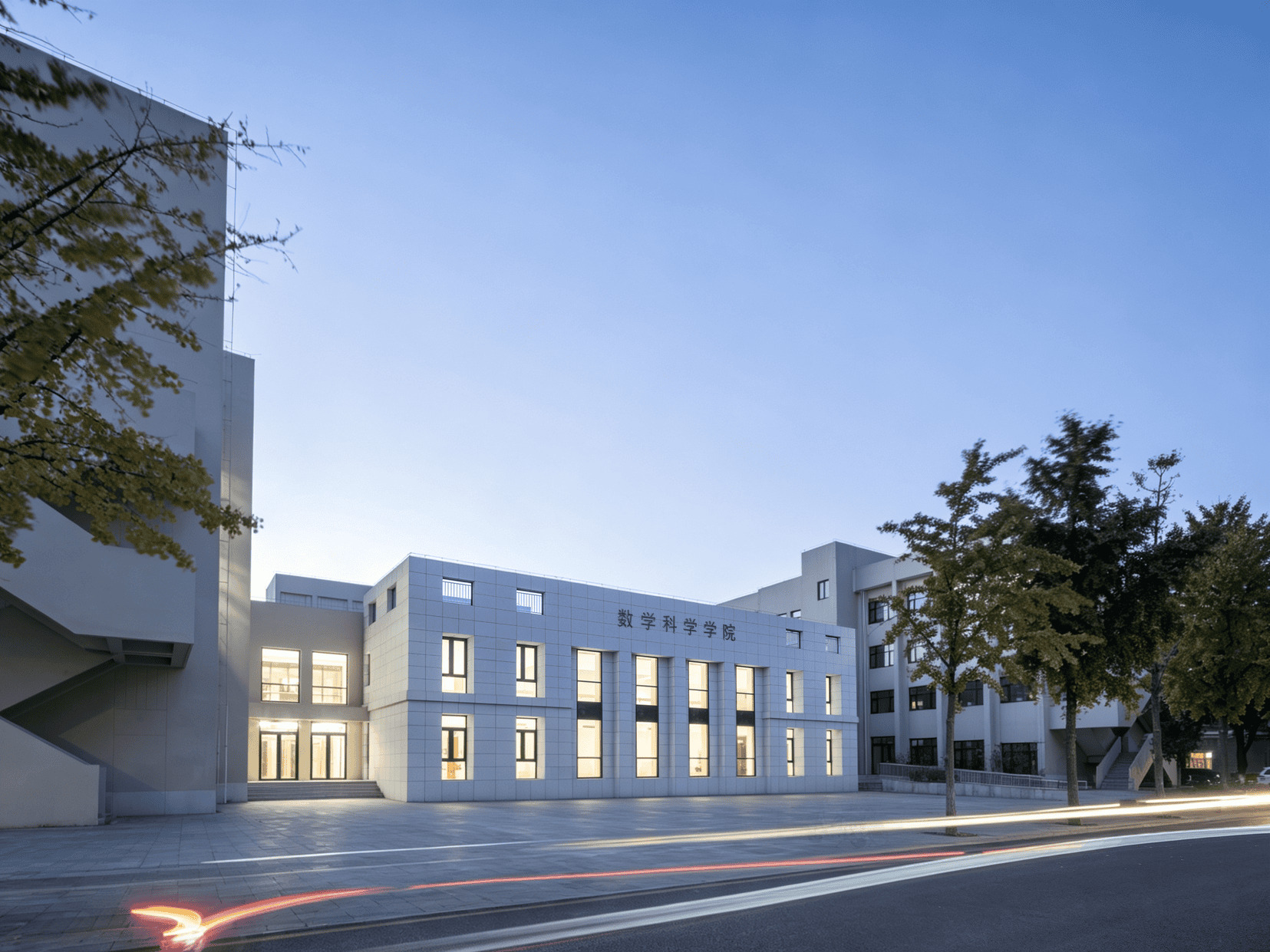 Pale tiled facade with vertical lit windows and rooftop signage at dusk, ginkgo trees and light trails from passing cars in the foreground