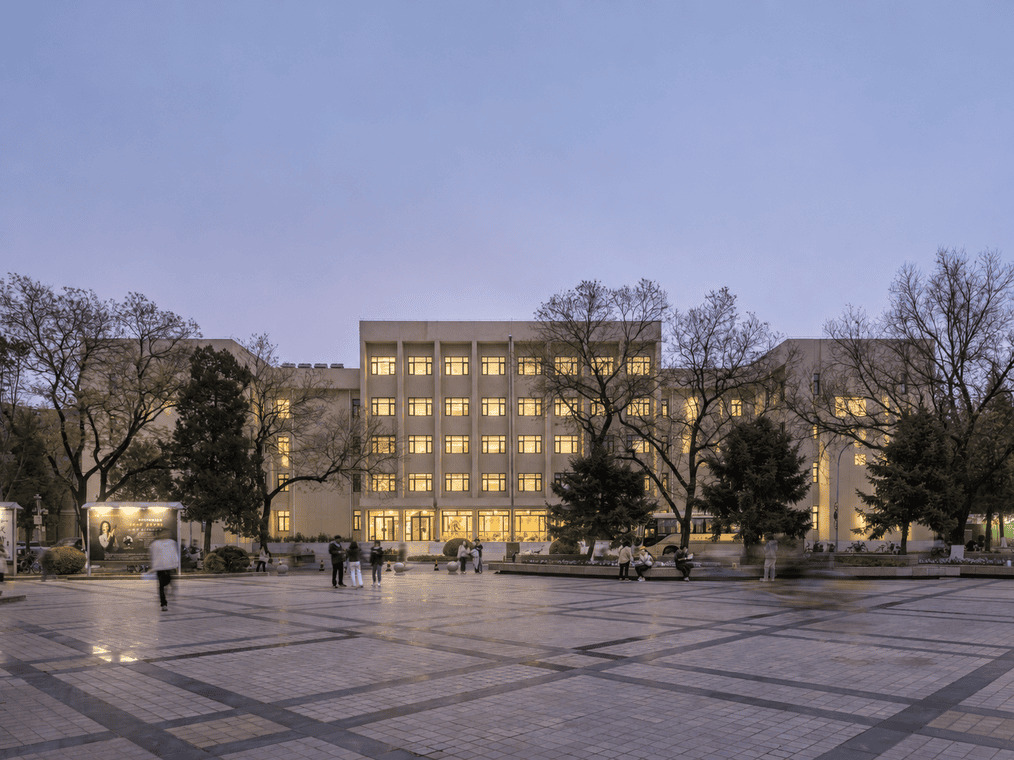 Five-storey building with lit windows beyond a patterned stone paving plaza, bare trees and a few people crossing at dusk