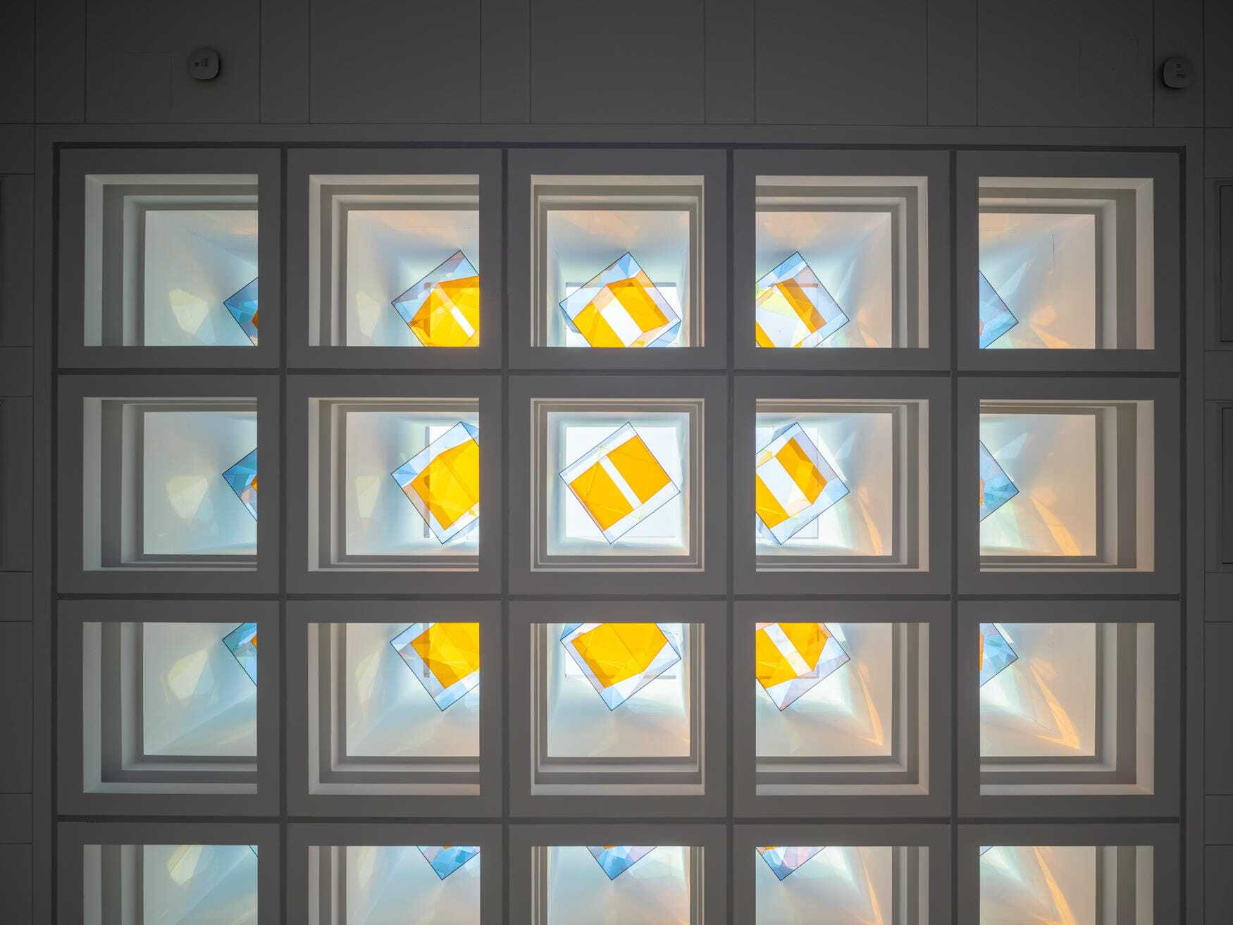 Looking up at a grid of square skylight coffers with yellow and blue tinted glass panels turned at angles