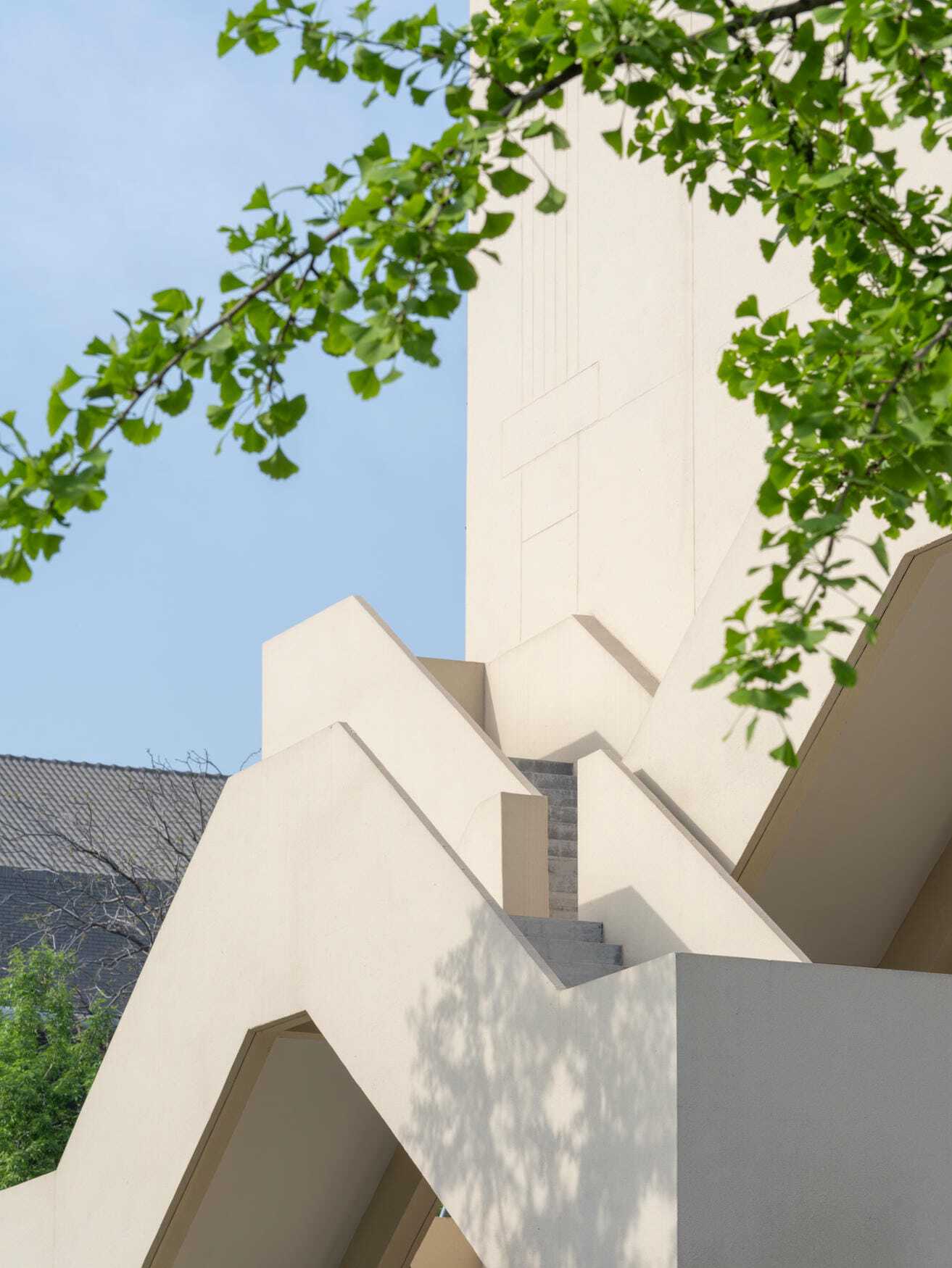 Close view of white concrete stair flights zigzagging up a tower wall, ginkgo leaves hanging across a blue sky