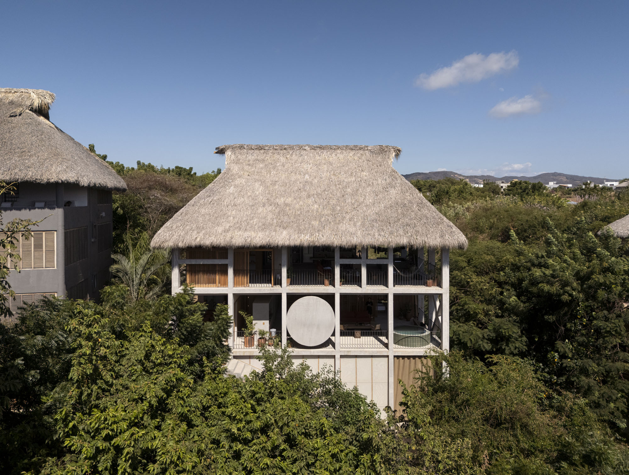 Frontal view of the thatched-roof house on a concrete frame with a round disc panel, rising above dense green canopy under a blue sky