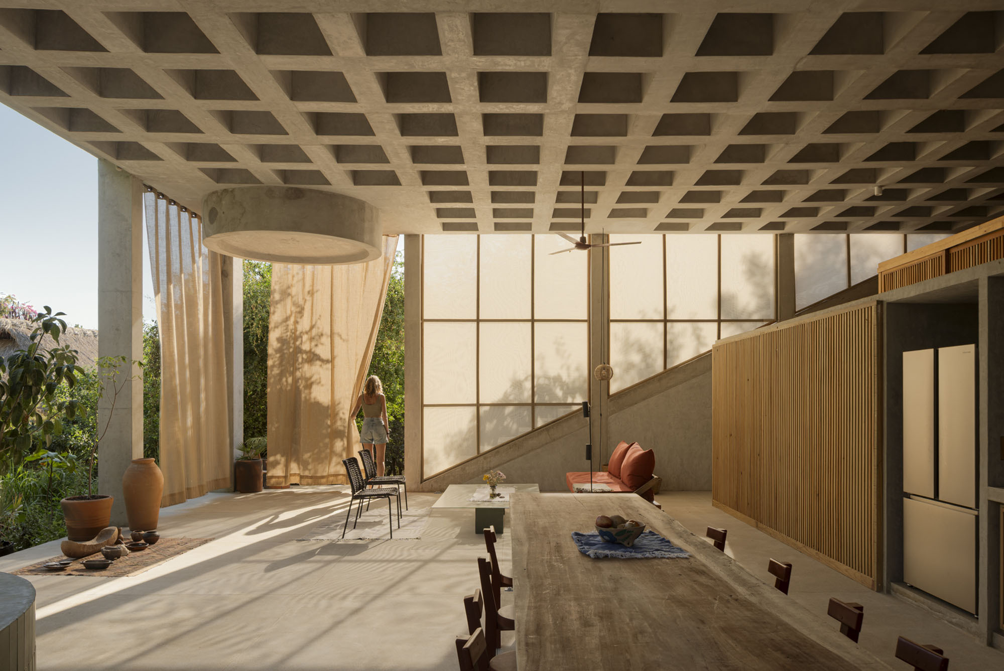 Open living space under a concrete waffle ceiling, with translucent panels, linen curtains and a woman standing in dappled sunlight