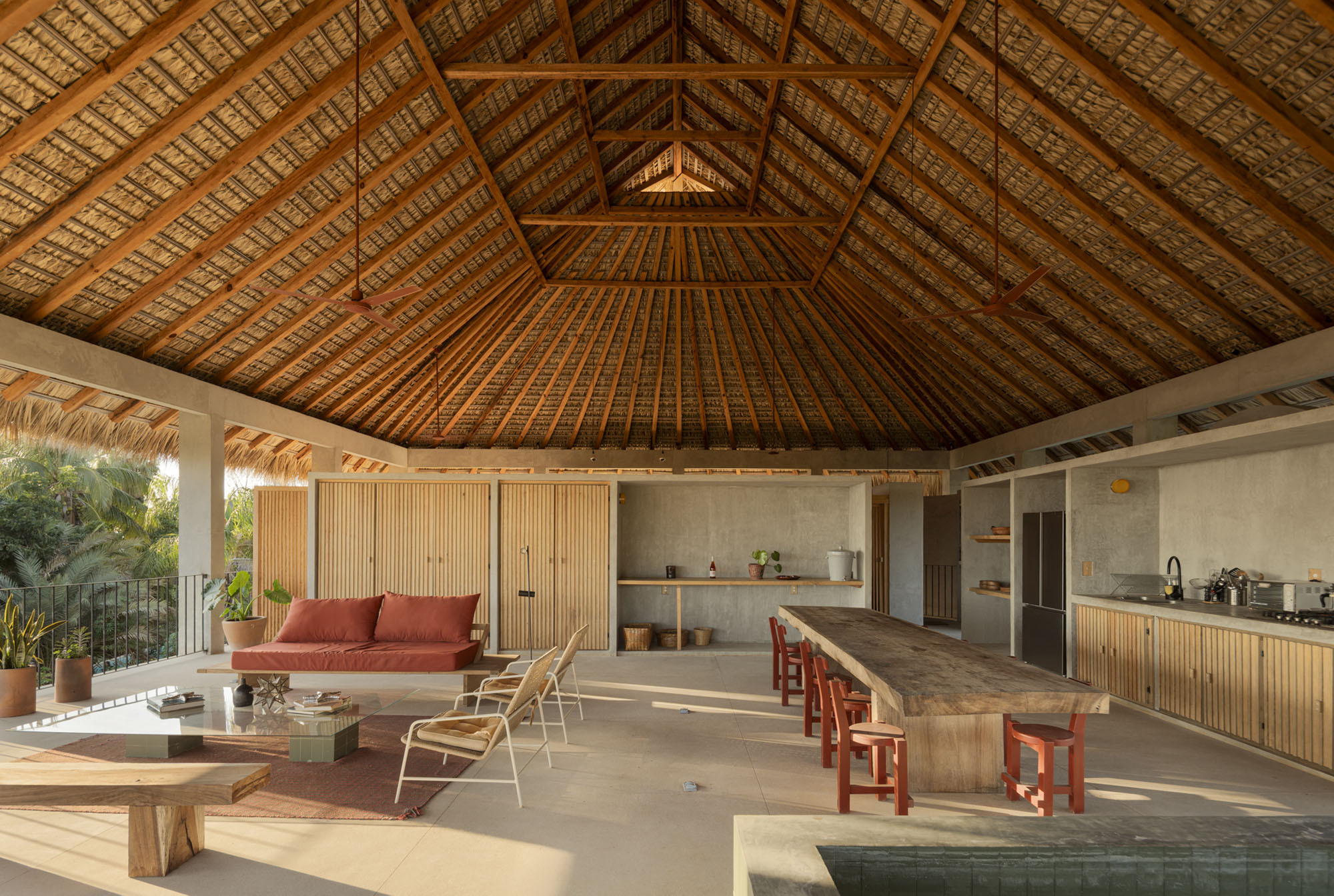 Interior under a pitched palm-thatch roof with timber rafters, a long wooden table, red stools and a red sofa in late light