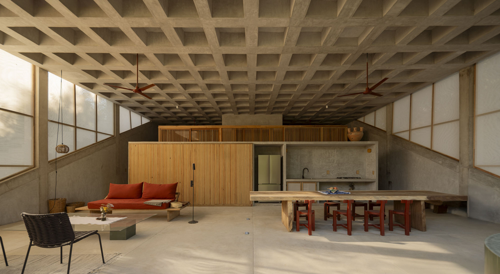 Concrete waffle-slab ceiling above an open room with slatted timber cabinetry, a red daybed, ceiling fans and a long dining table