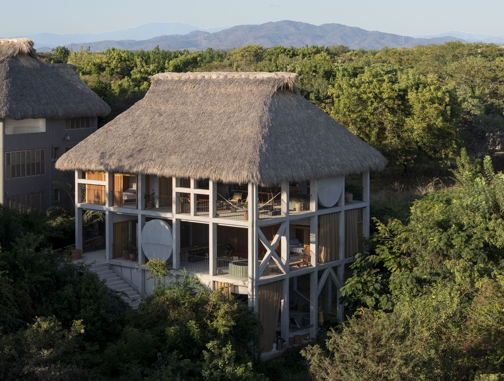 Elevated view of the thatched house with its white concrete frame, X-shaped bracing and round disc panels among green trees at golden hour