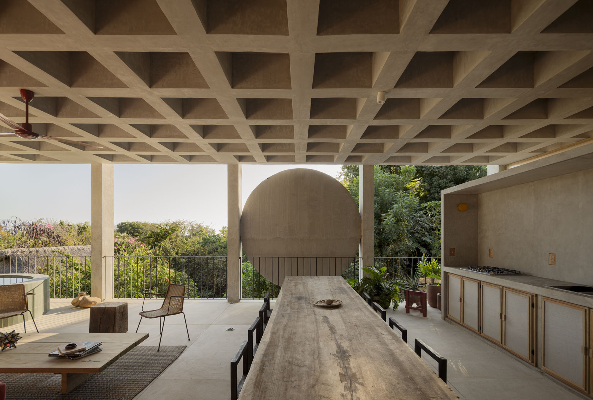Open terrace under a concrete waffle-slab ceiling, with a long timber table facing a round wall disc and leafy treetops