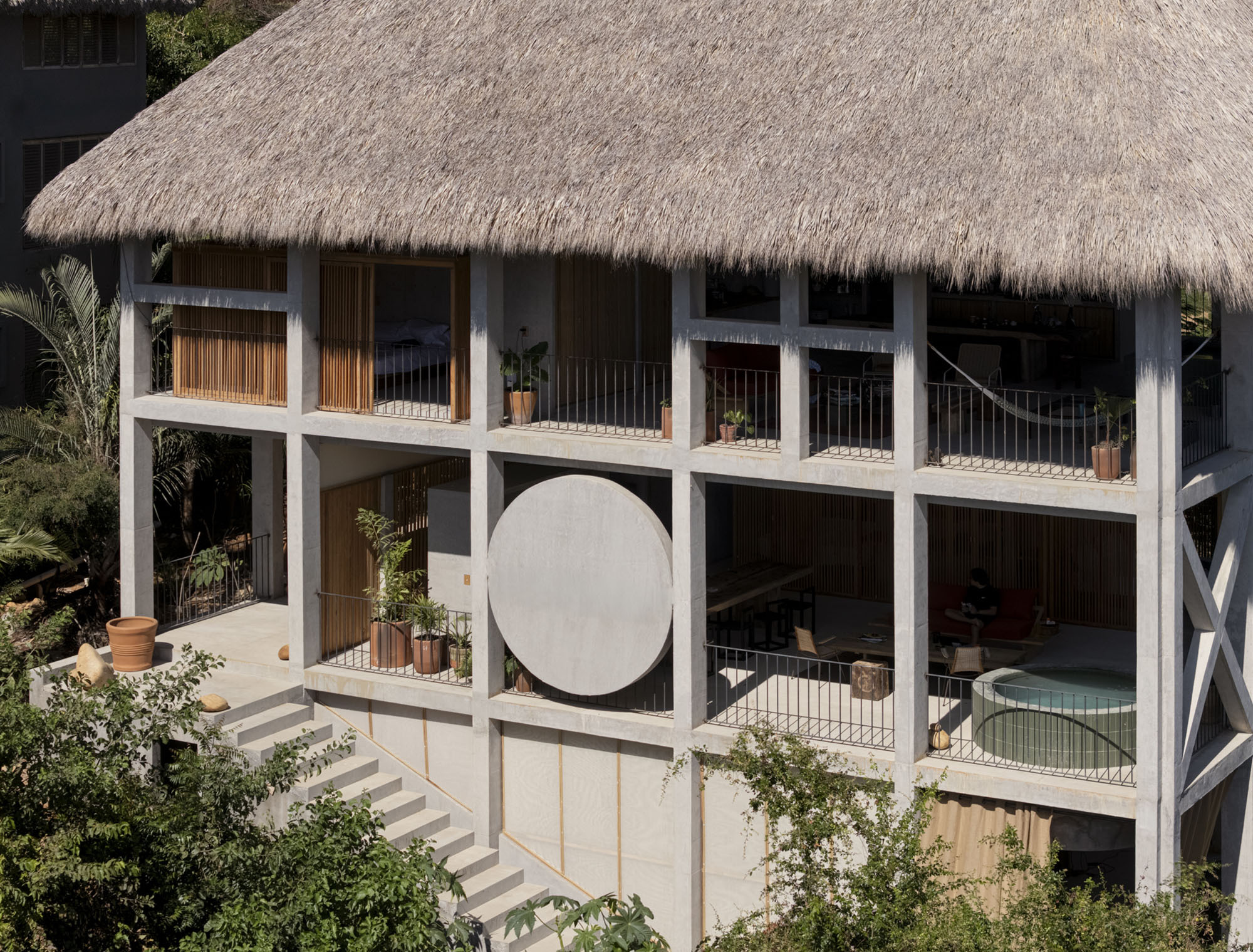 Exposed concrete frame with a thatched roof, round wall disc, balconies, plunge pool and stone stair among tropical foliage