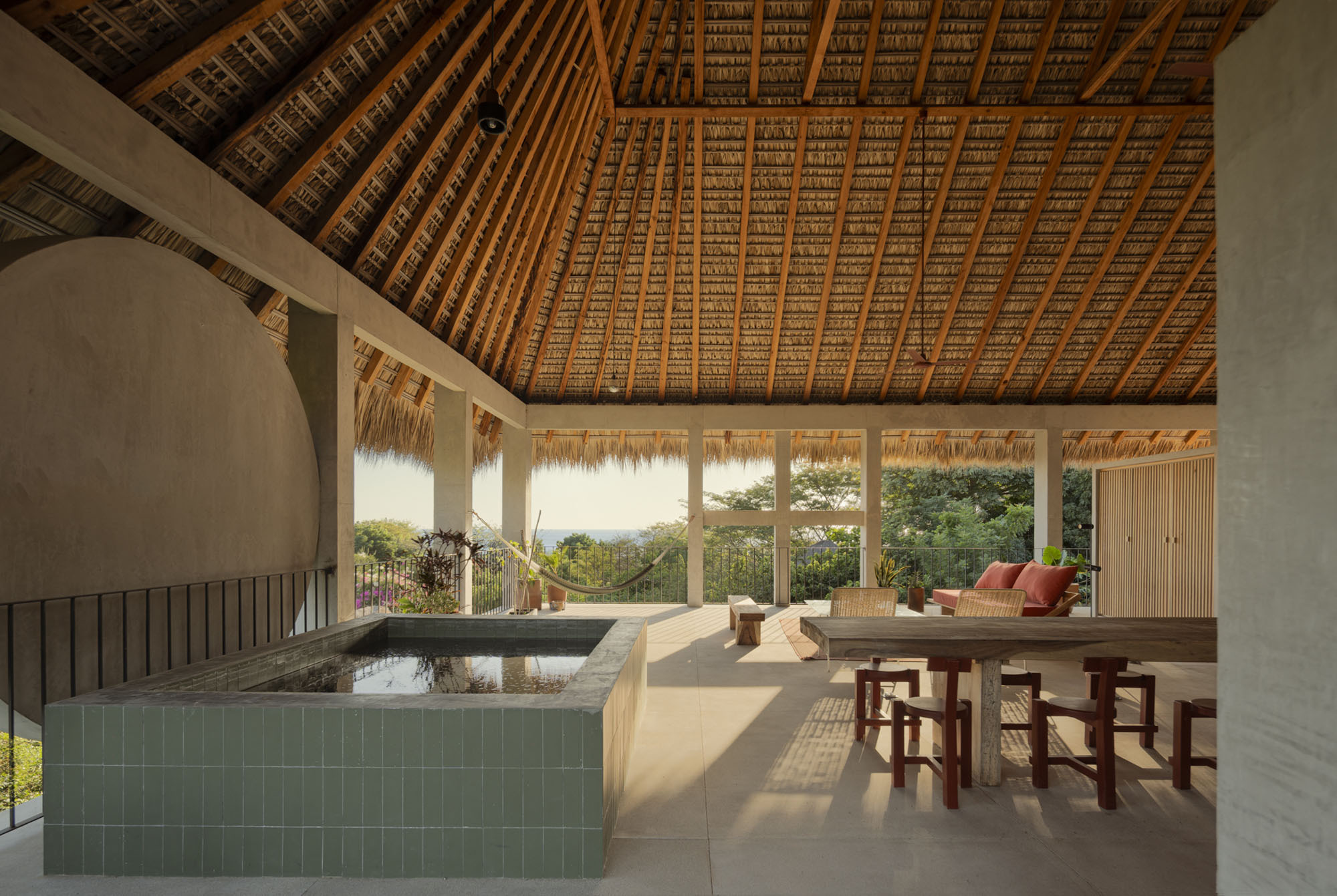 Terrace beneath a pitched palm-thatch ceiling, with a green tiled plunge pool, wooden dining table and hammock in dappled light