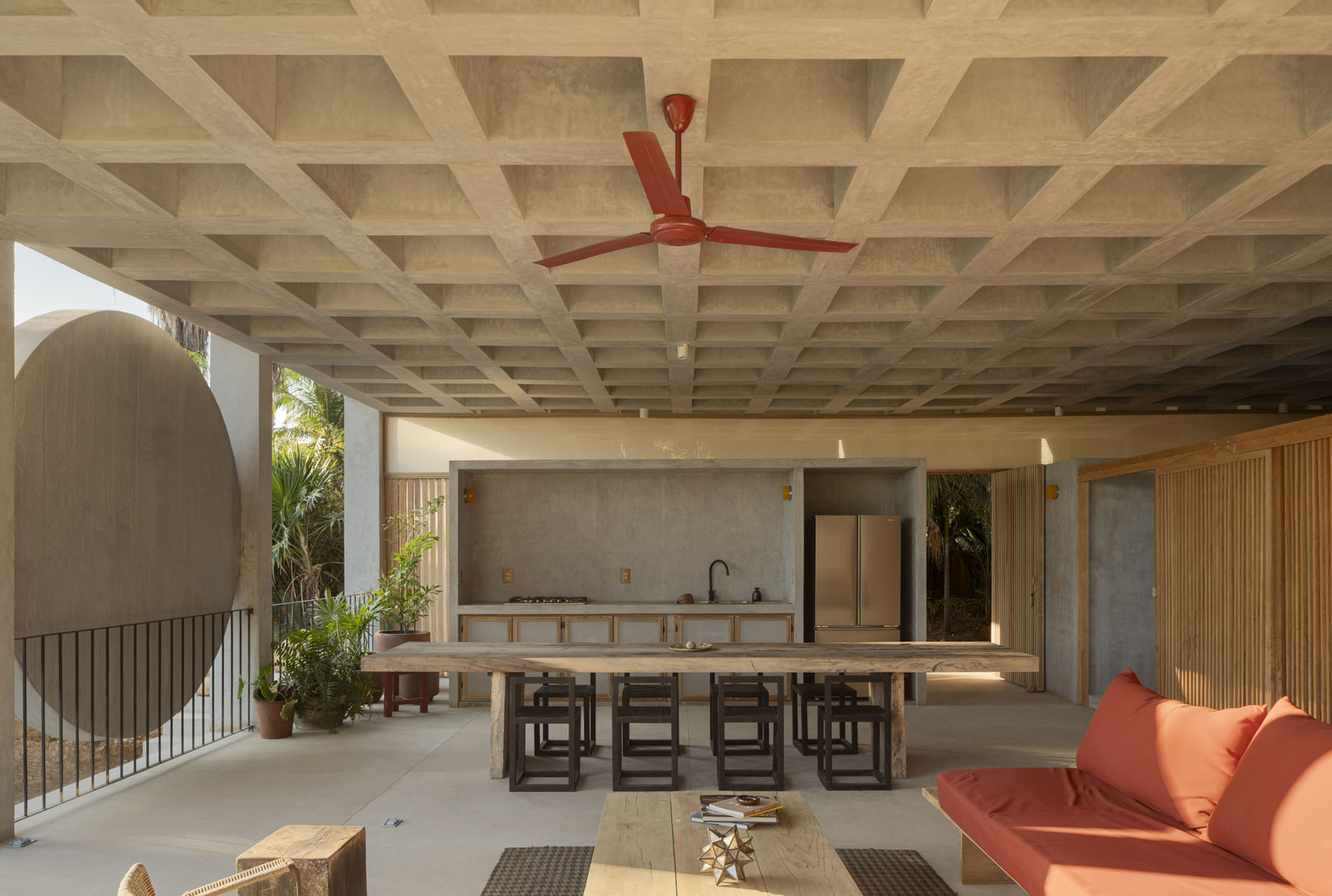 Open-air kitchen and dining area under a coffered concrete ceiling, with a red ceiling fan and coral cushioned daybed