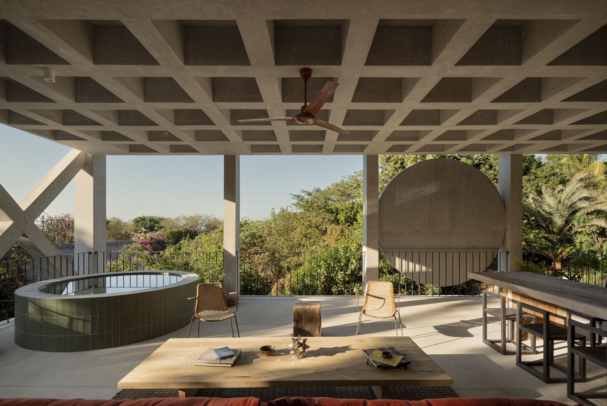 Terrace under a coffered concrete ceiling, with a round green-tiled plunge pool, low timber table and cane chairs in afternoon sun