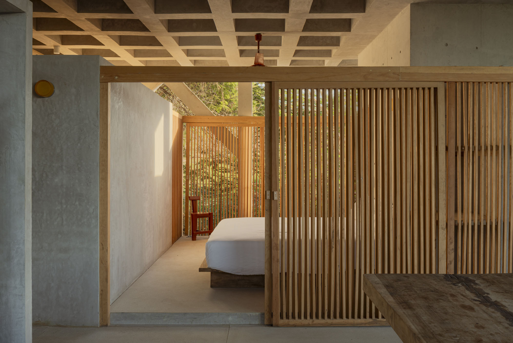 Bedroom alcove behind a sliding timber slat screen, with a waffle concrete ceiling and a red chair in dappled afternoon light