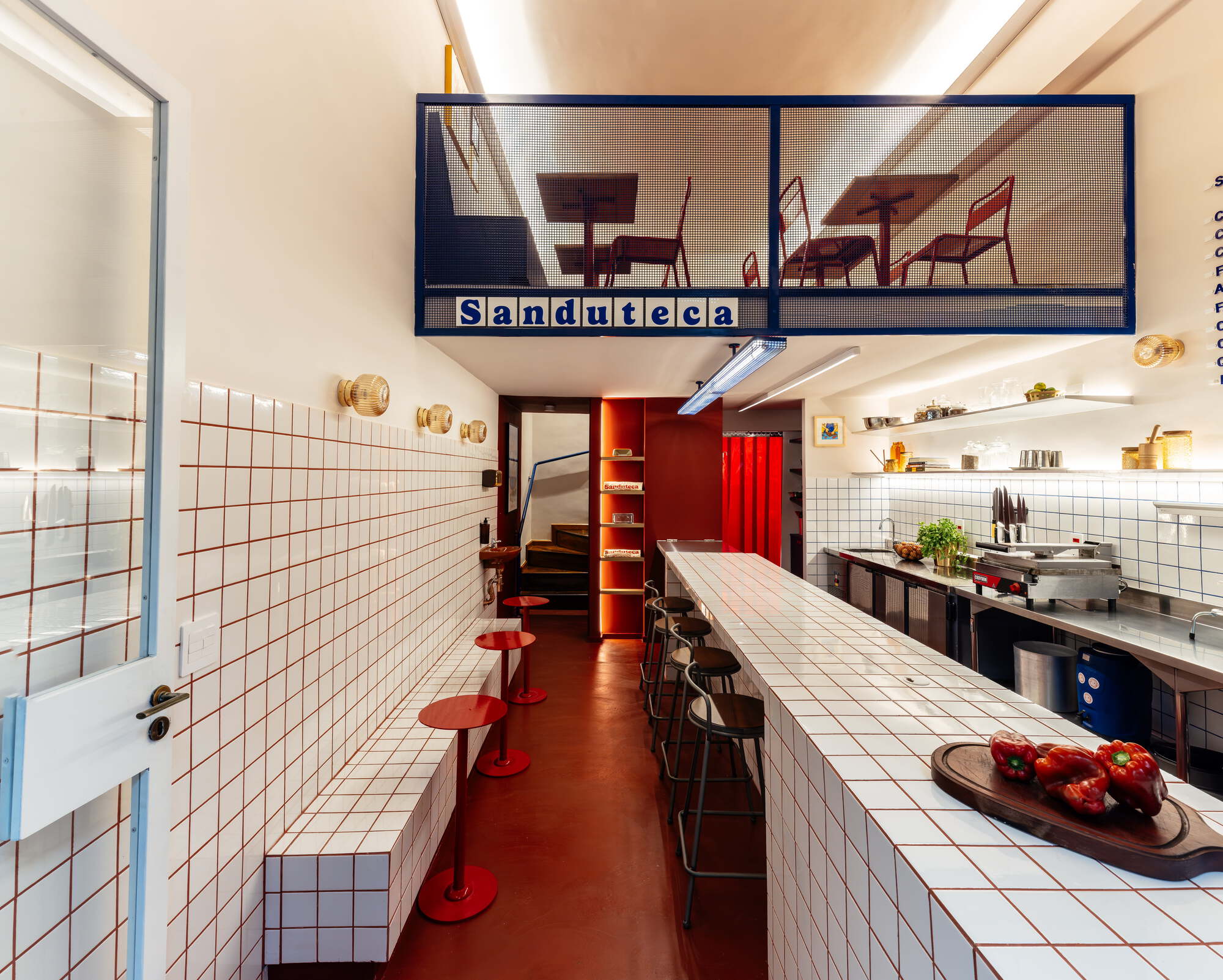 Narrow sandwich shop interior with white grid-tiled counter, red stools and a blue mesh mezzanine, lit by warm wall sconces and linear lights