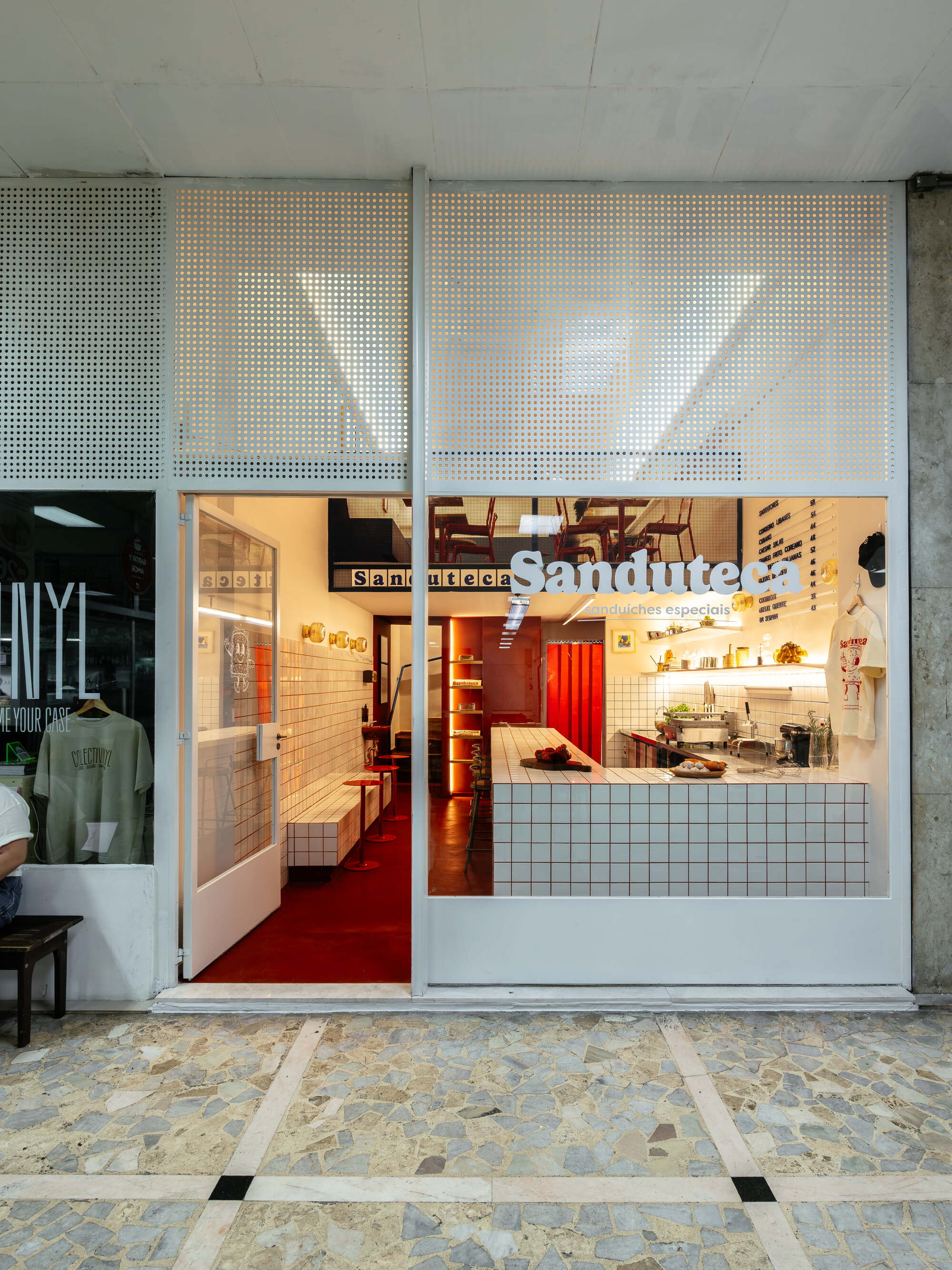 Shopfront seen from the patterned stone pavement, with perforated white metal panels above glazing that reveals the red-floored interior