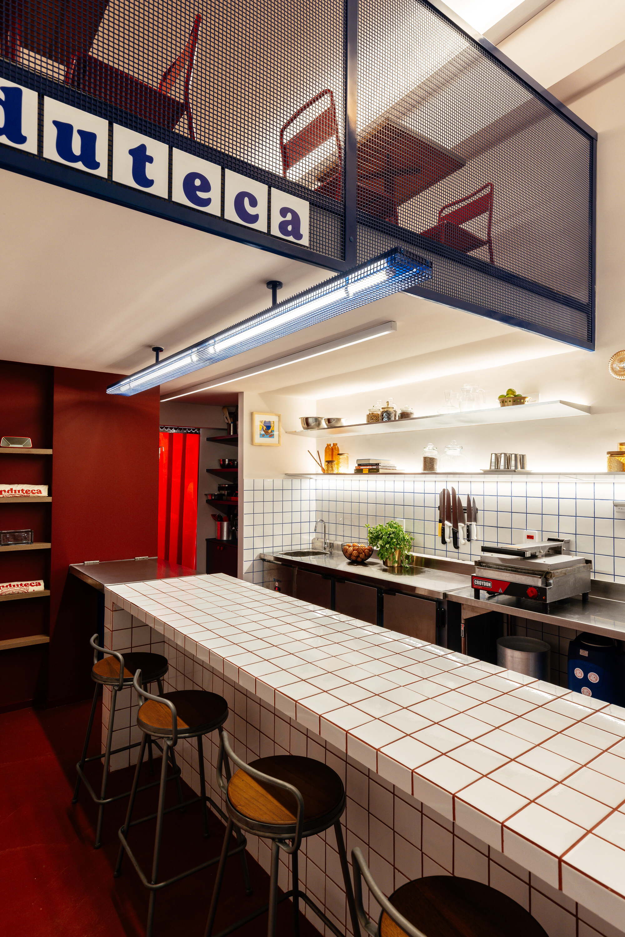 White tiled counter with red grout and wooden stools below a blue mesh mezzanine, lit by a long fluorescent tube