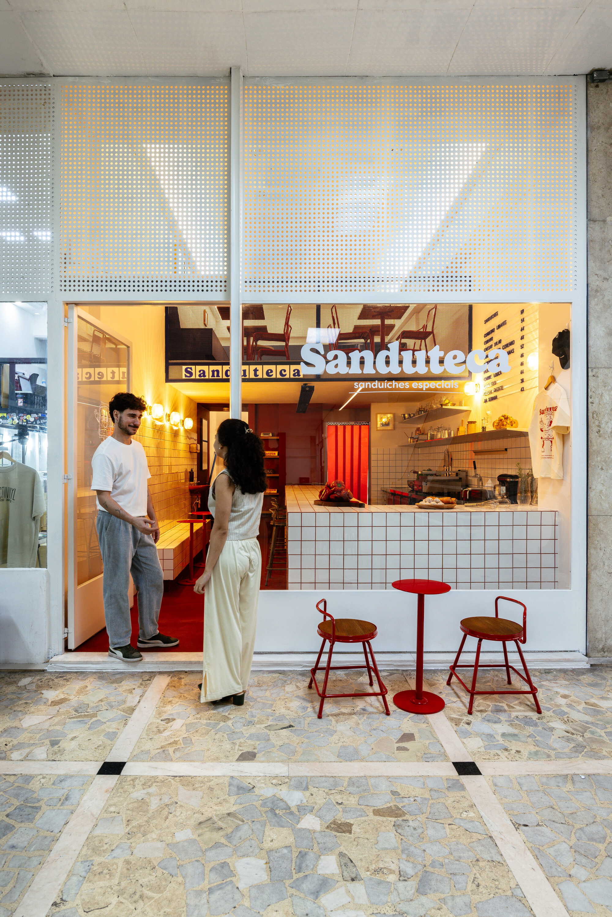 Storefront with perforated white metal panel above a tiled counter, red stools and two people talking at the open door