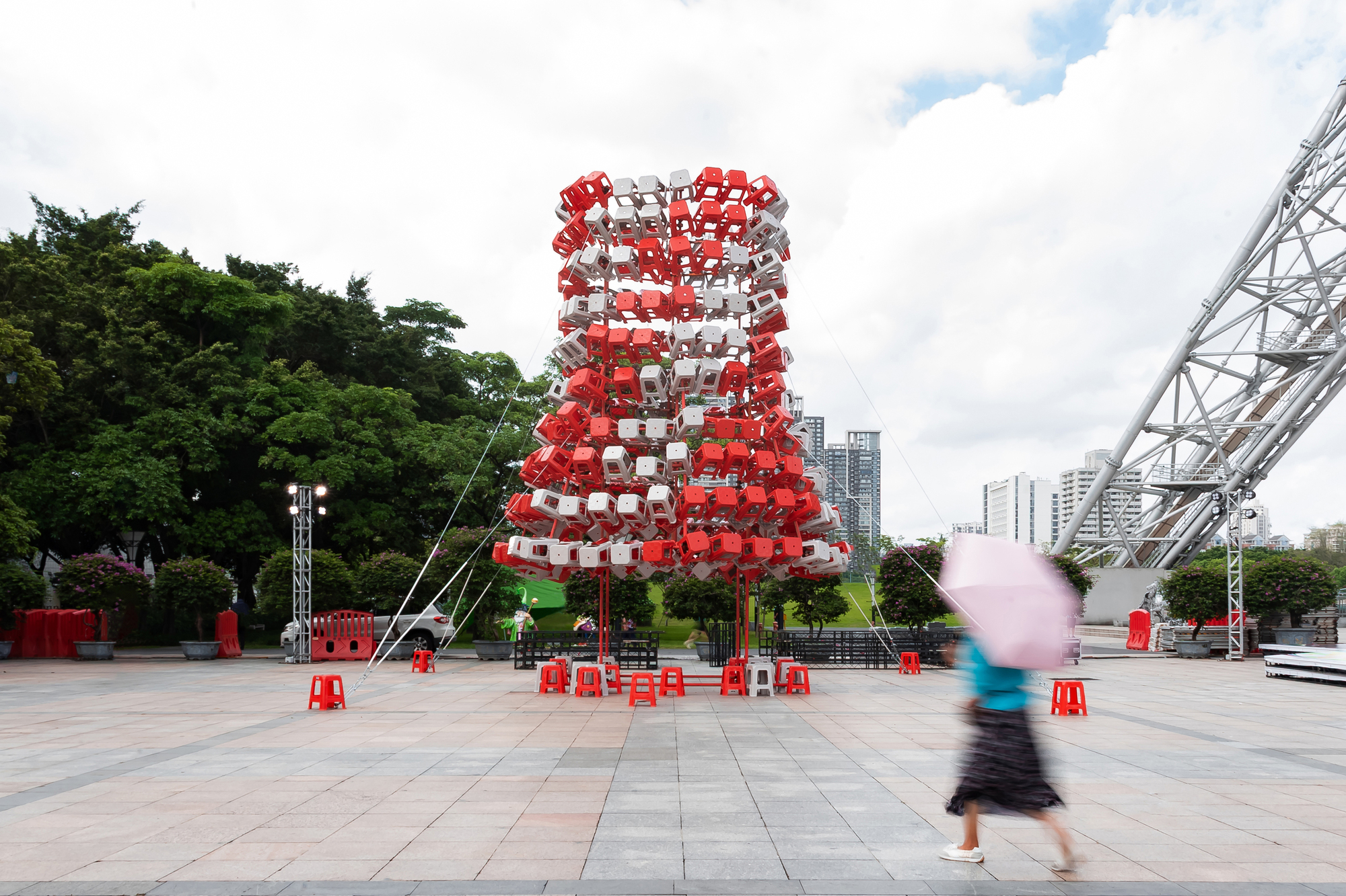 Cylindrical tower of stacked red and white plastic stools on a paved plaza, with a blurred passerby holding a pink umbrella