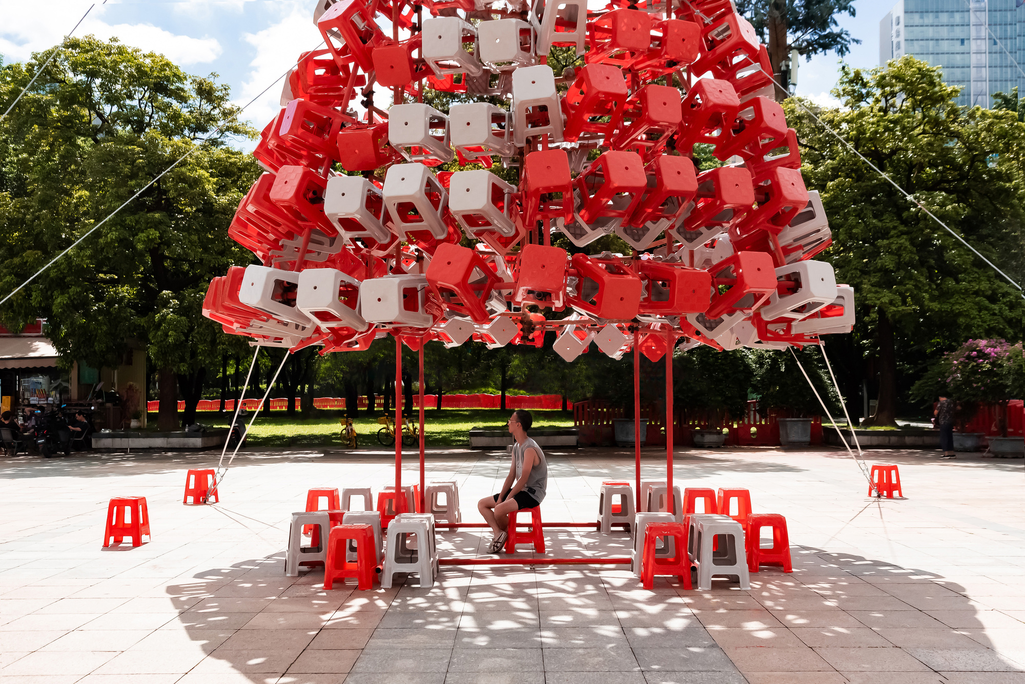Seated visitor beneath the canopy of red and white stools, with dappled tree shadows across the plaza paving