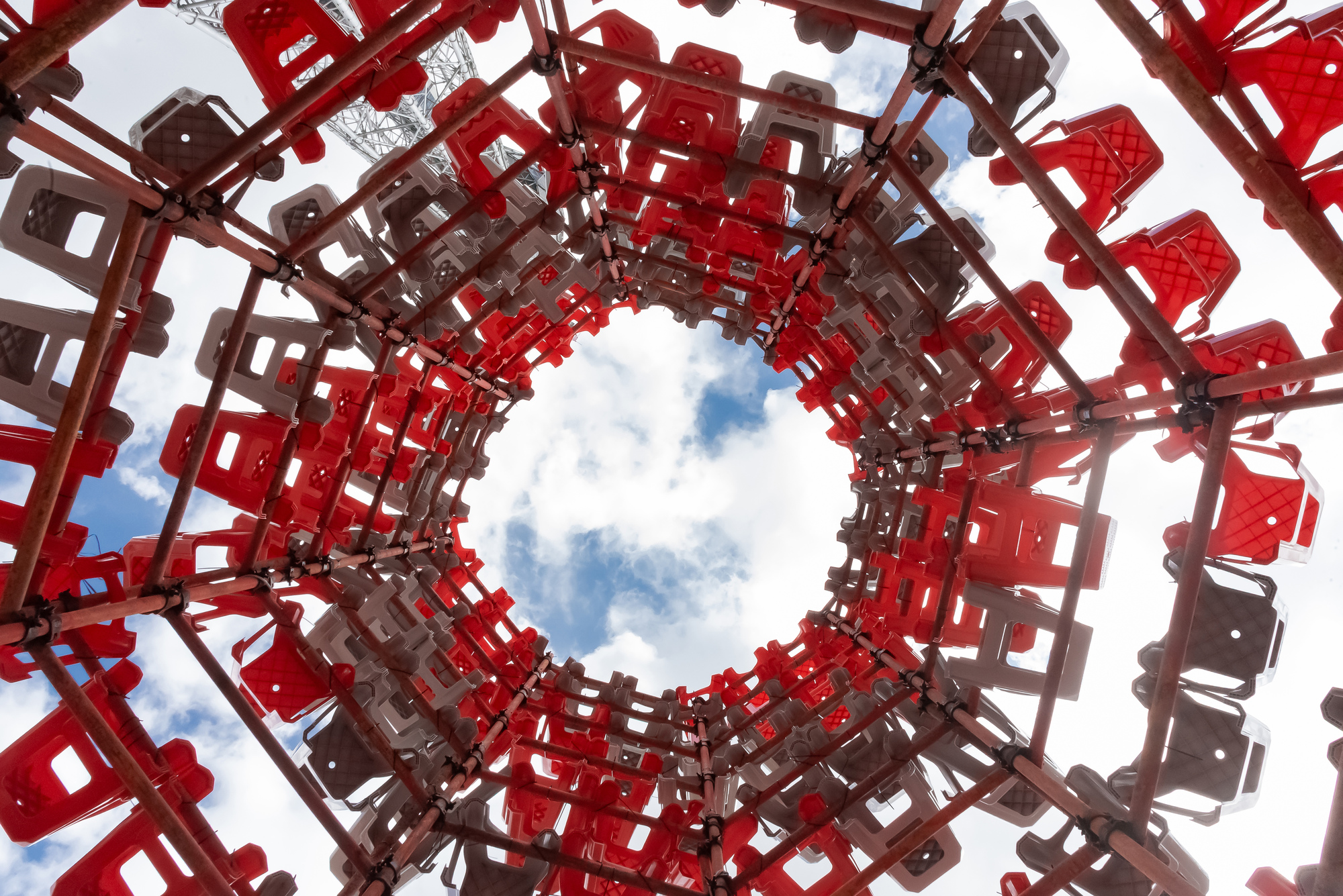 Upward view through the open center of the stool structure, with steel tube clamps framing a cloudy sky