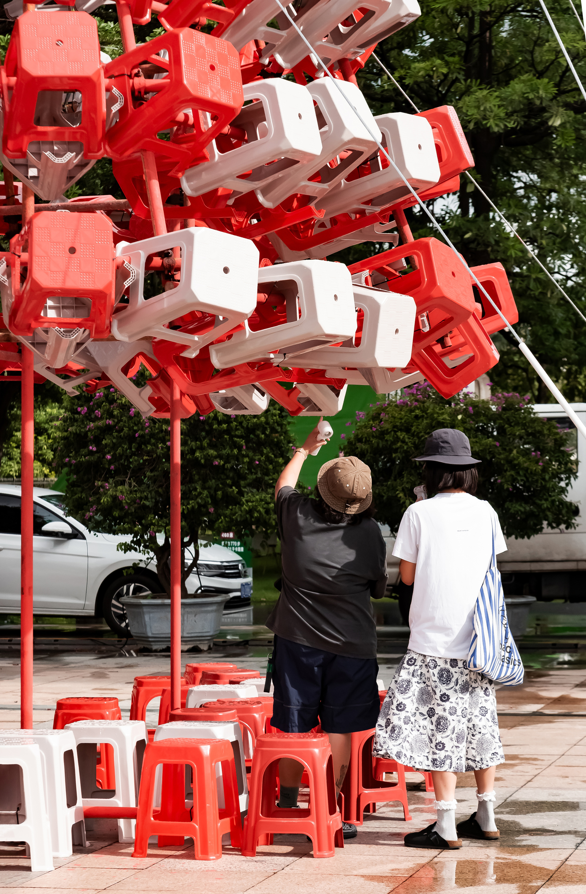 Two visitors on the wet plaza, one reaching up to touch the underside of a white stool in the cluster