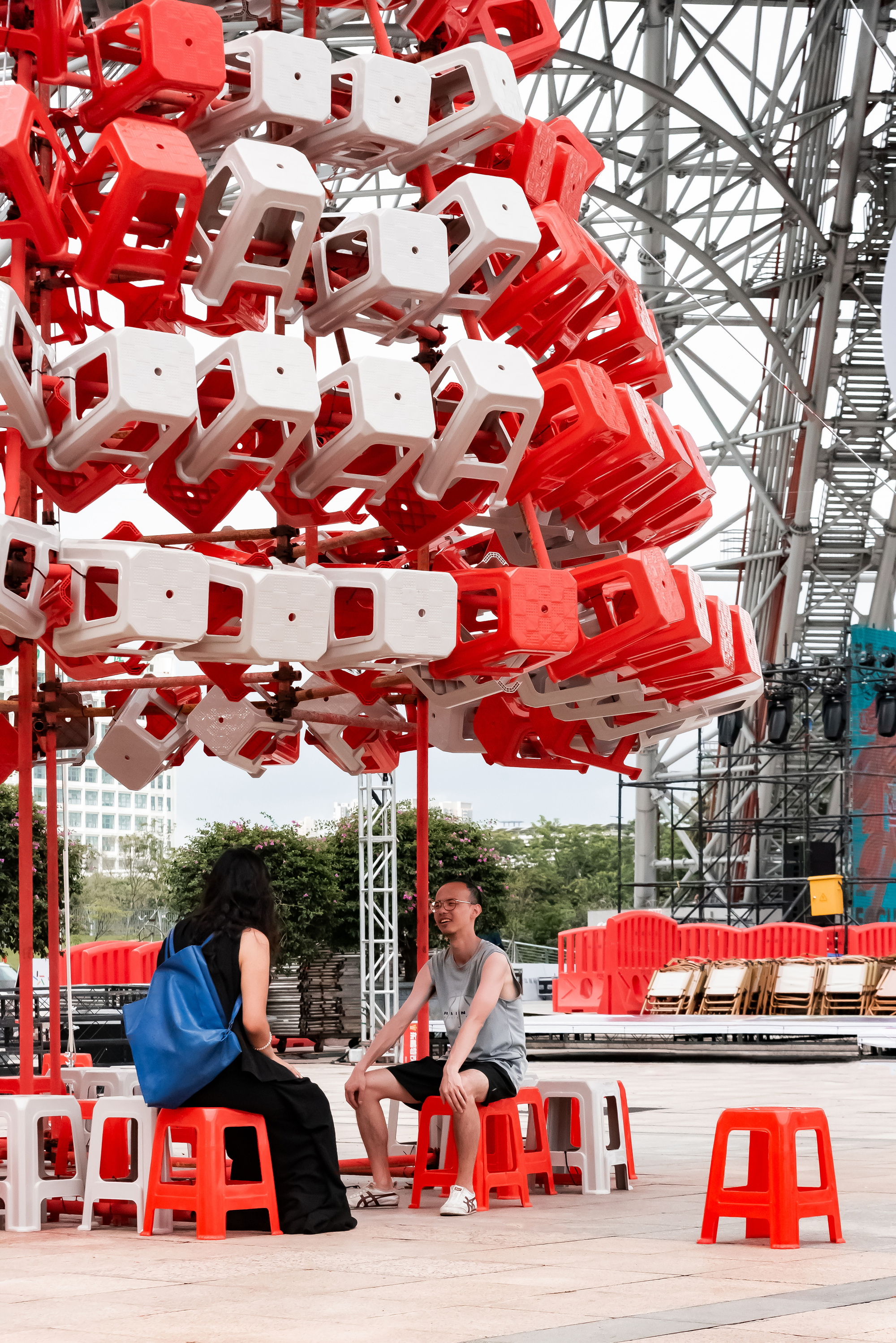 Two people seated on low red stools under the hanging stool canopy, with a steel truss stage behind