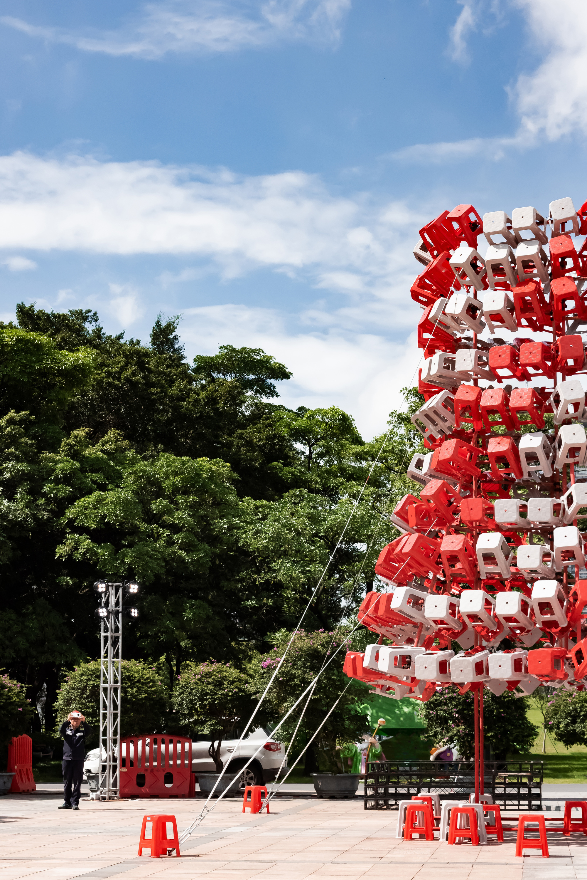 Tower of stacked red and white plastic stools held by guy lines on a paved plaza under blue sky