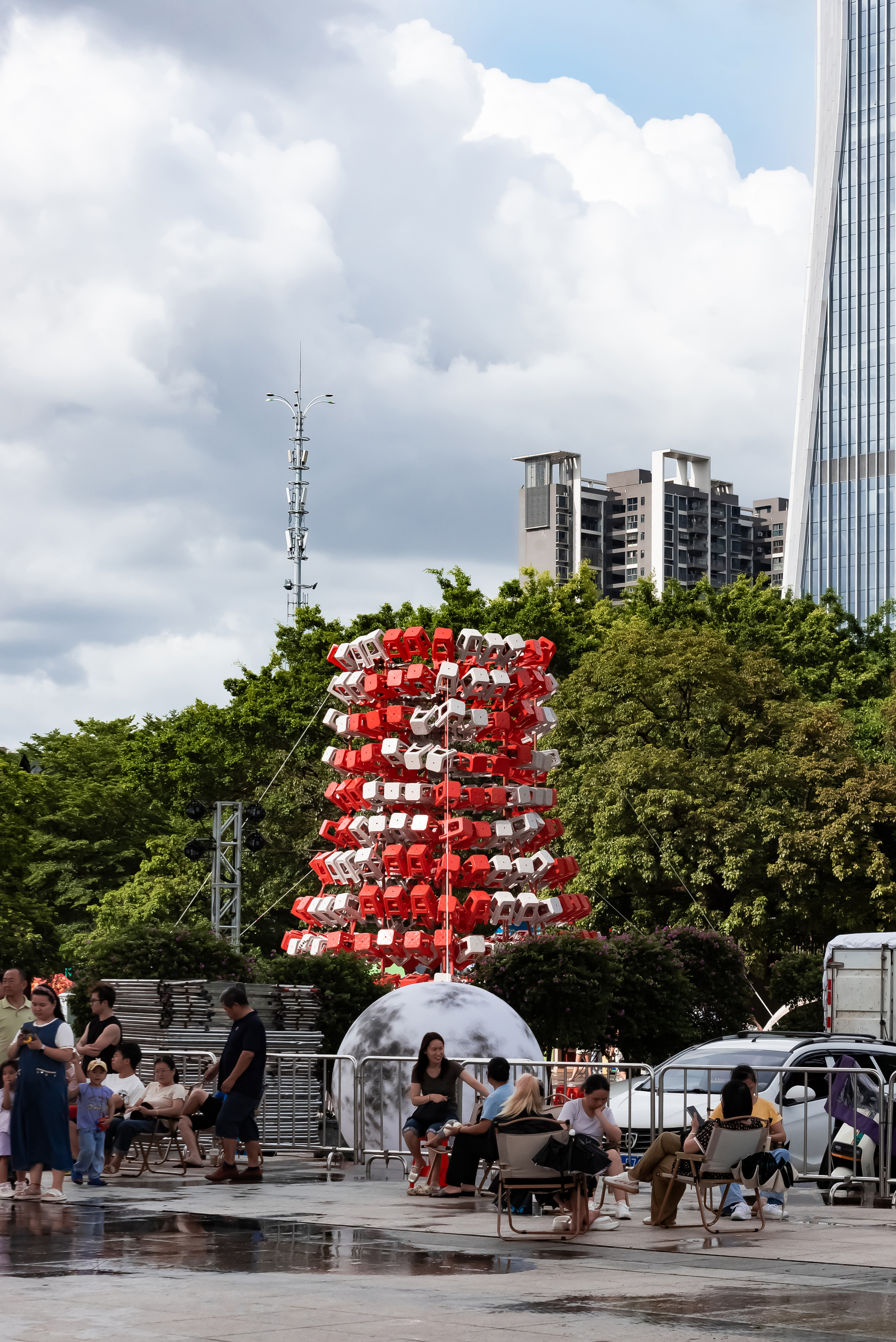 Distant view of the stool tower rising behind seated visitors on a wet plaza under heavy clouds