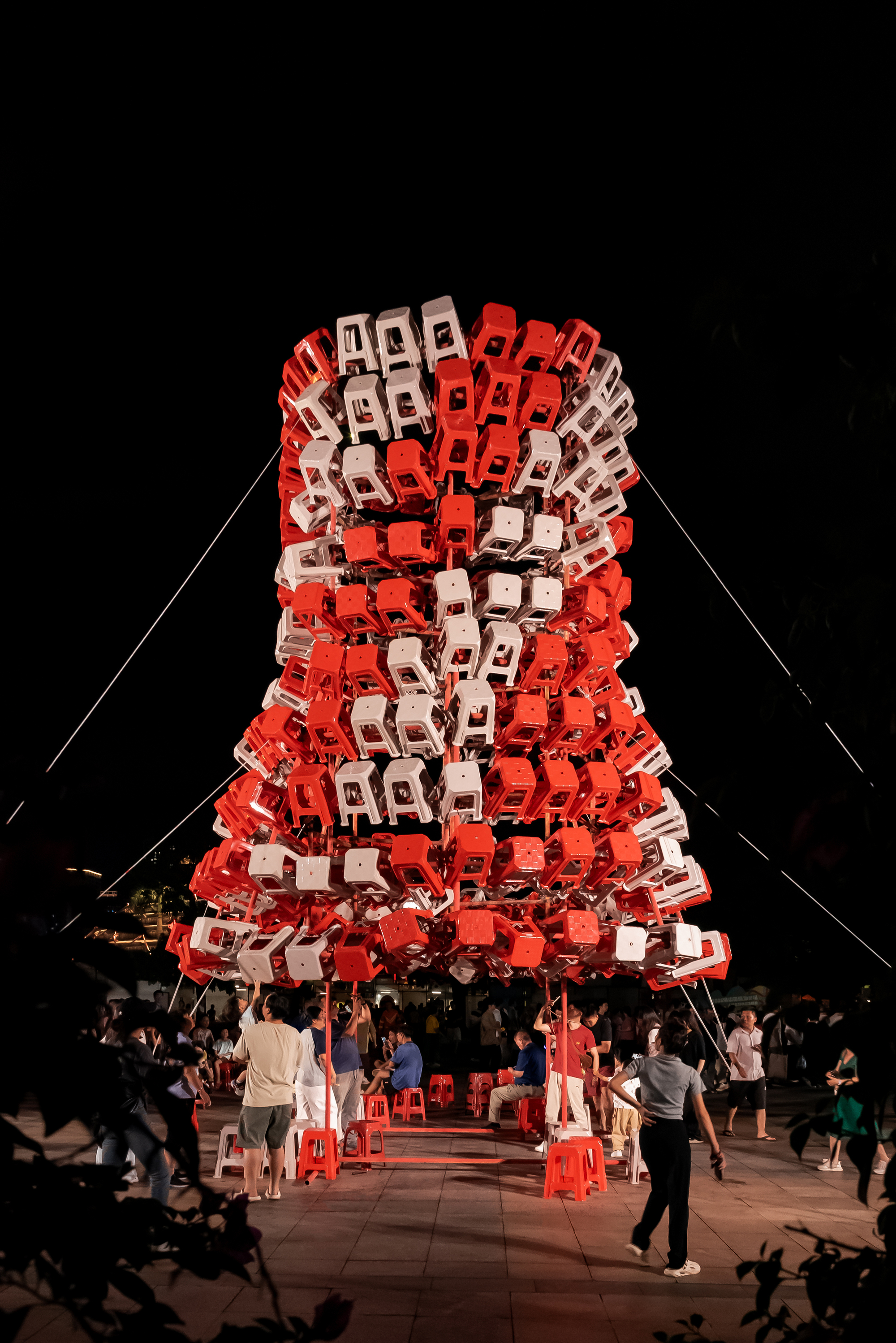 Night view of the stool tower glowing red and white as people gather and sit beneath it